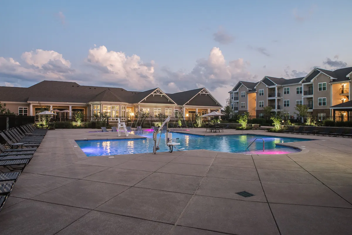 Charming evening view of an upscale apartment complex with an inviting illuminated pool and comfortable lounge chairs, perfect for relaxation and leisure.