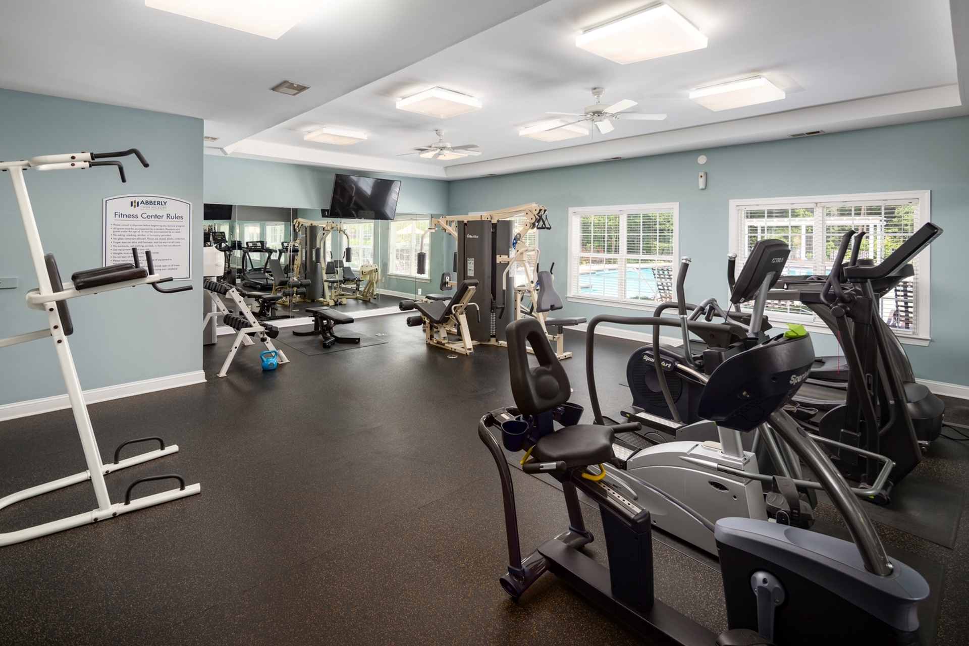Modern fitness center with state-of-the-art equipment, large mirrors, and ample natural light from wide windows overlooking a pool area.
