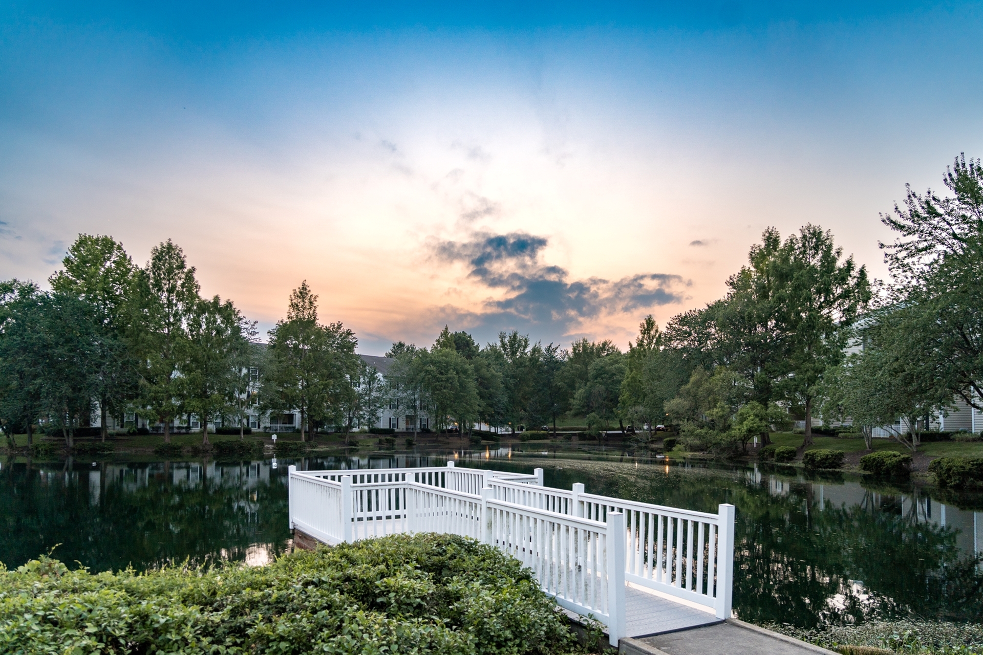 Stunning sunset view over a serene lake with lush green trees, a charming white bridge, and elegant apartment buildings in the background.