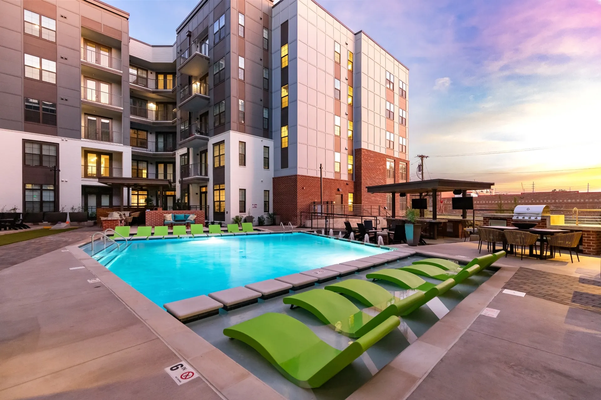 Luxurious apartment complex featuring a sparkling outdoor pool with stylish green loungers and a welcoming outdoor entertainment area at sunset.