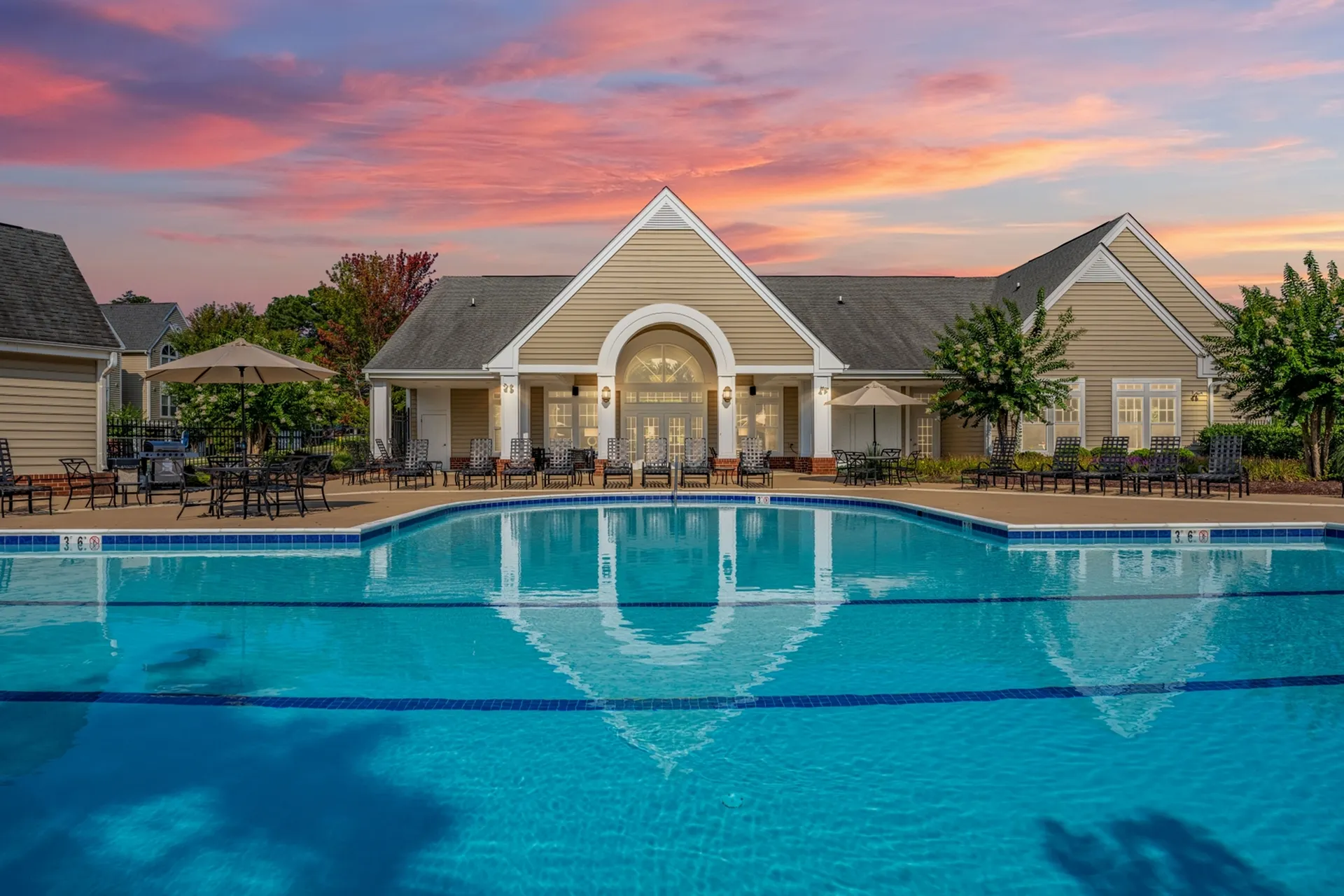 Luxurious clubhouse with a large swimming pool, surrounded by stylish outdoor seating and lush landscaping, under a stunning sunset sky.