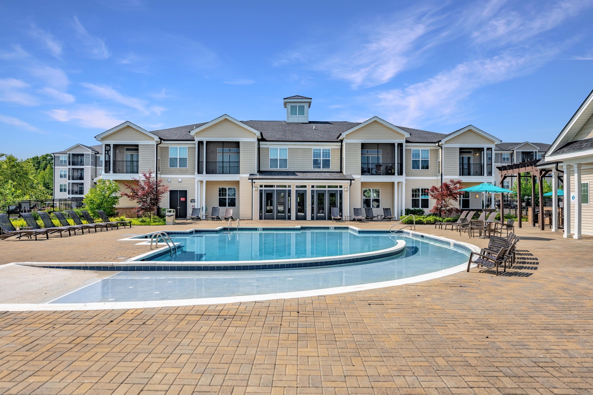 Elegant apartment complex with a stunning outdoor pool, surrounded by stylish lounge chairs and vibrant landscaping under a bright blue sky.