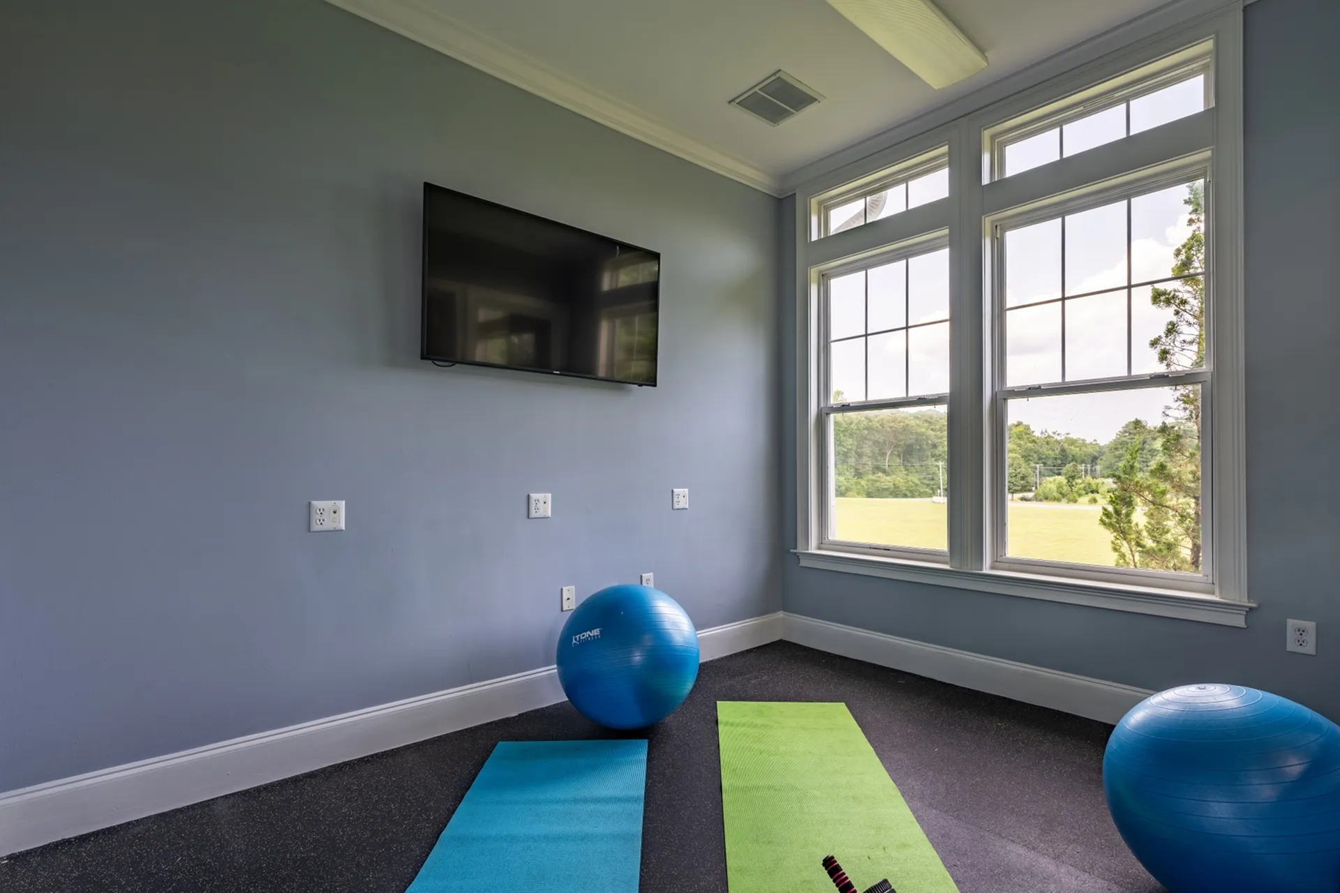 Bright exercise room with large windows, yoga mats, workout ball, and wall-mounted TV, overlooking a scenic view.