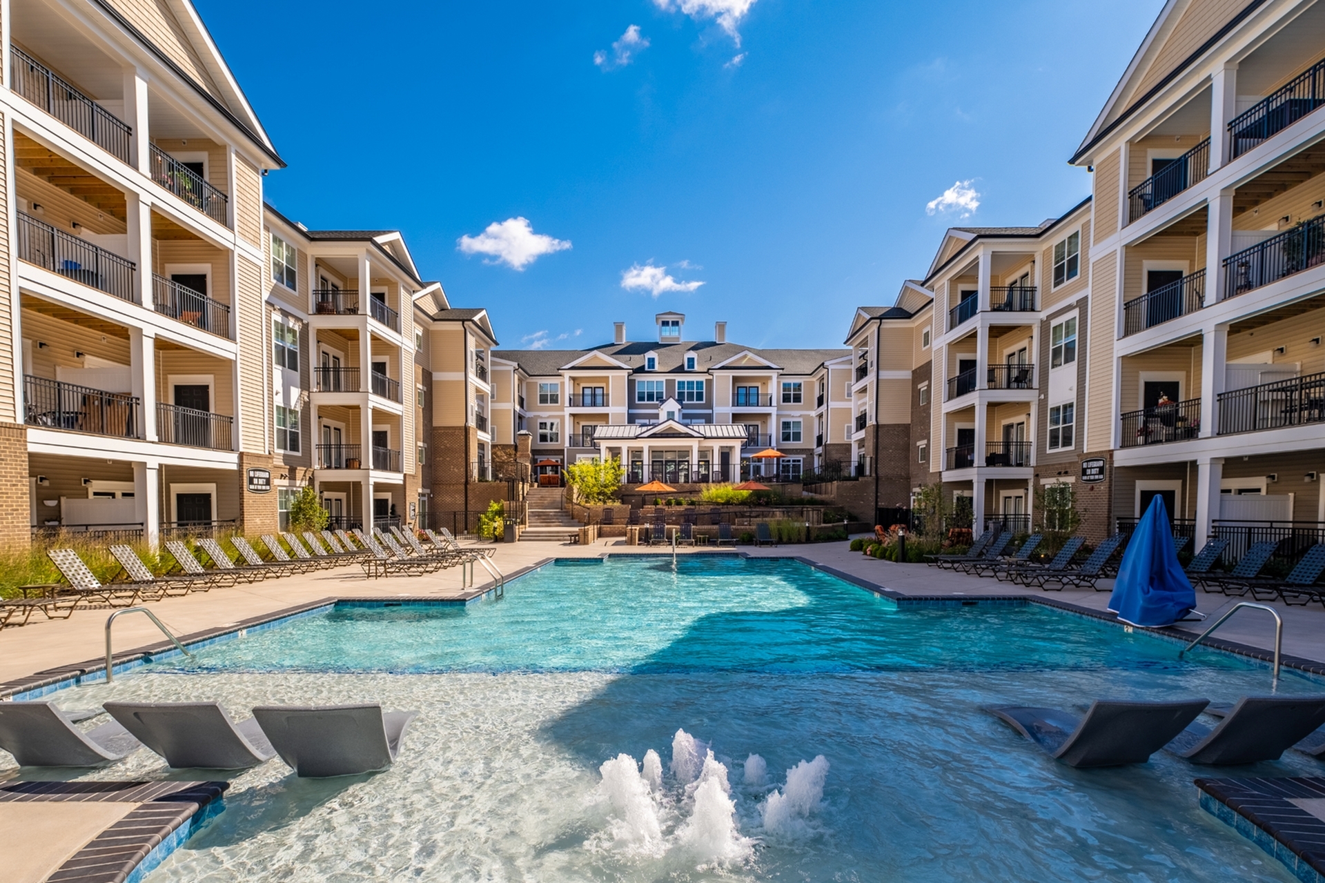 Luxurious apartment complex featuring a sparkling resort-style pool surrounded by comfortable loungers and elegant multi-story buildings under a clear blue sky.