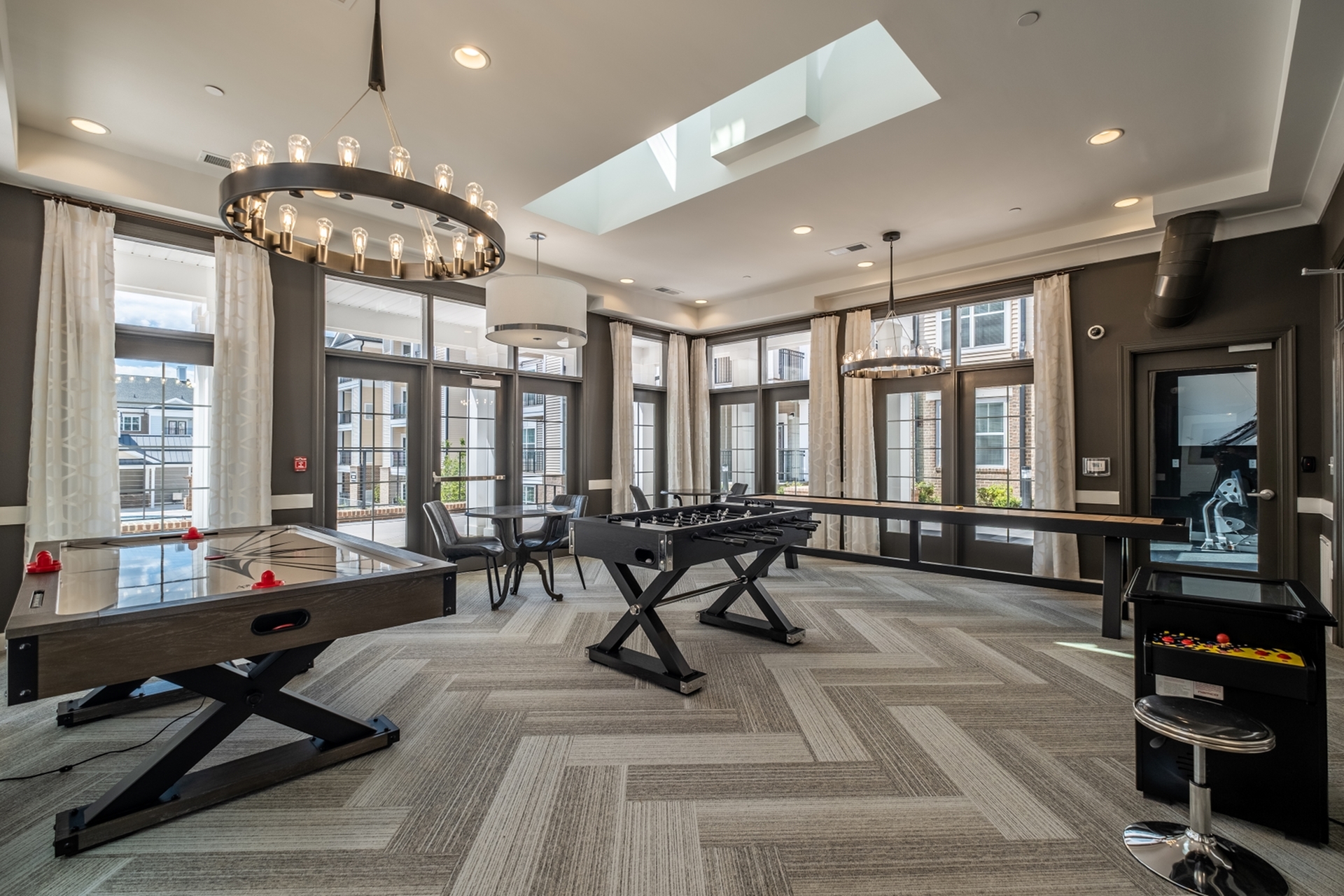 Spacious recreational room with modern foosball and air hockey tables, elegant chandeliers, and expansive windows offering abundant natural light.