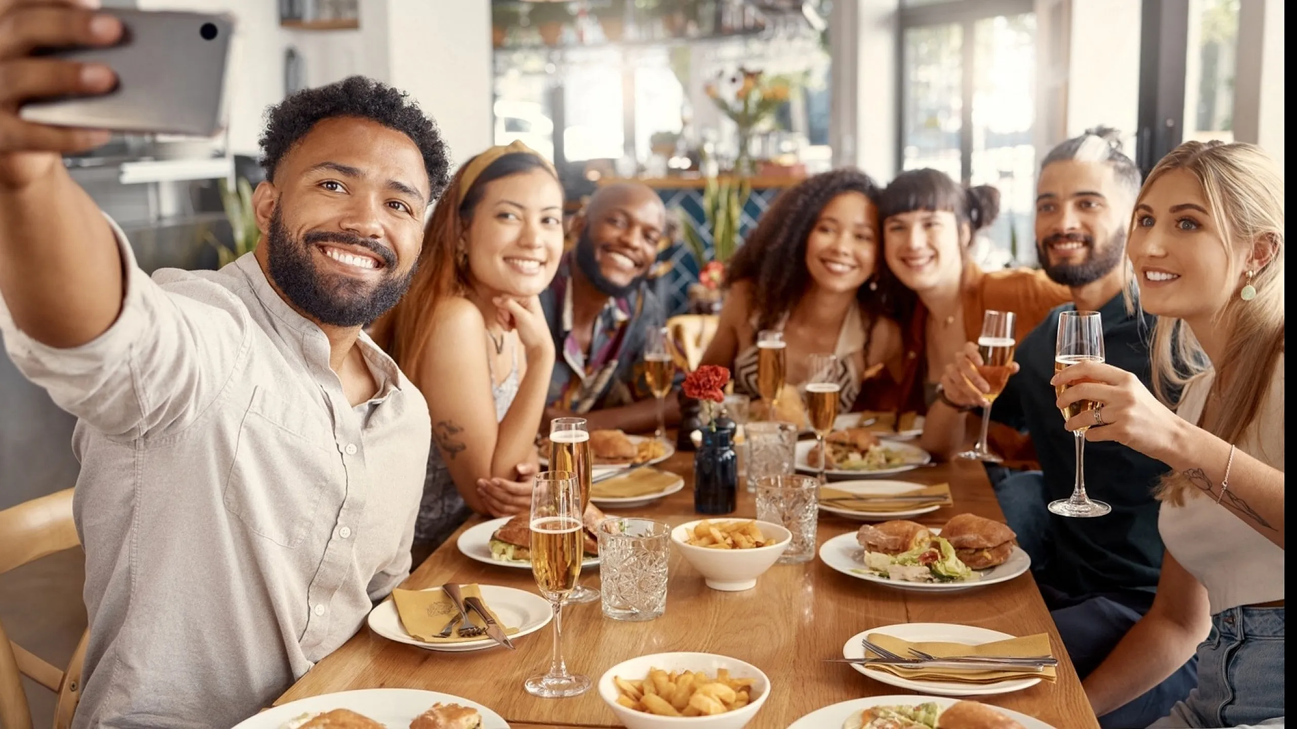 A group of friends at a restaurant table taking a selfie, with plates of food and drinks in front of them.