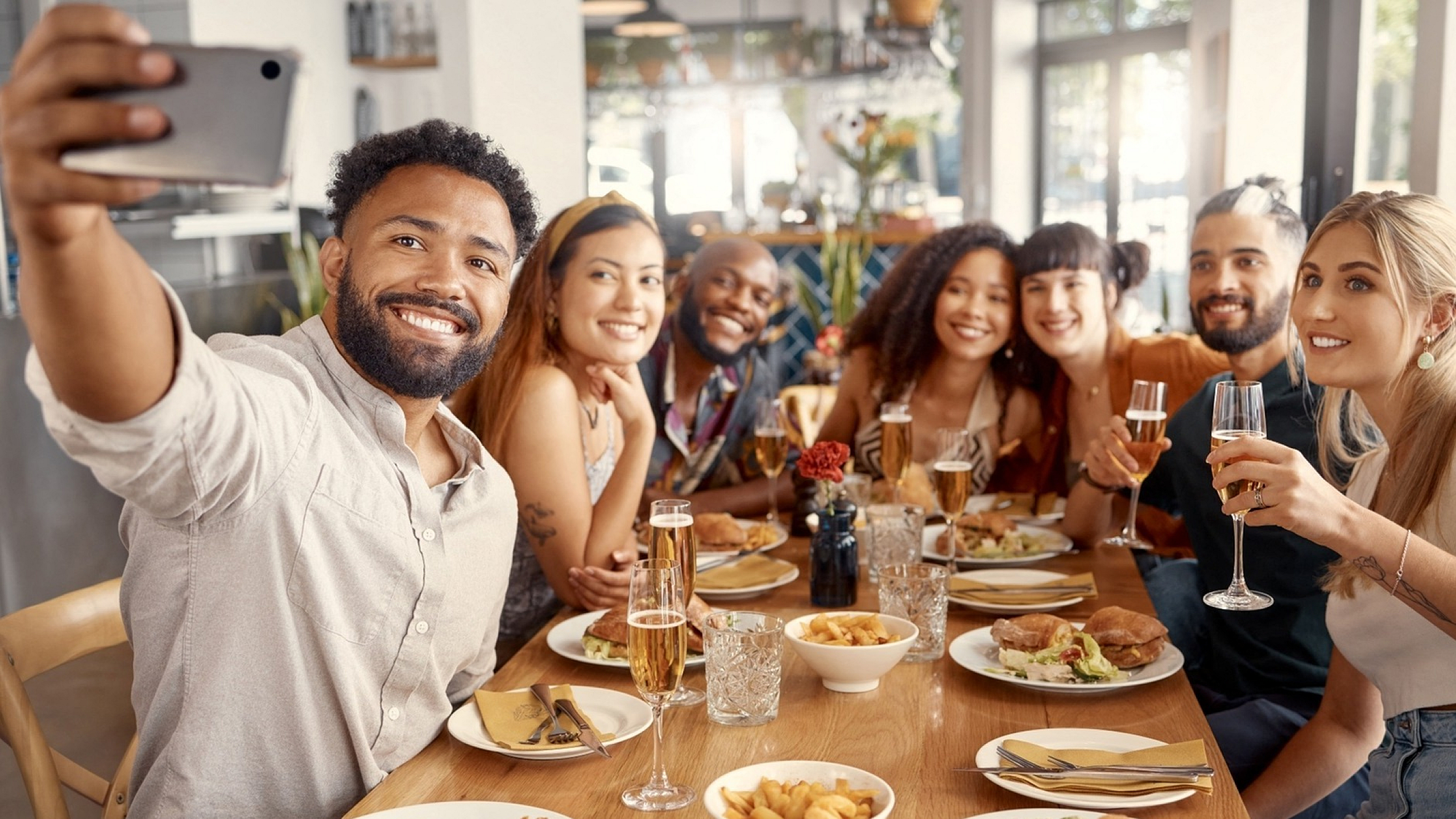 A group of friends at a restaurant table taking a selfie, with plates of food and drinks in front of them.