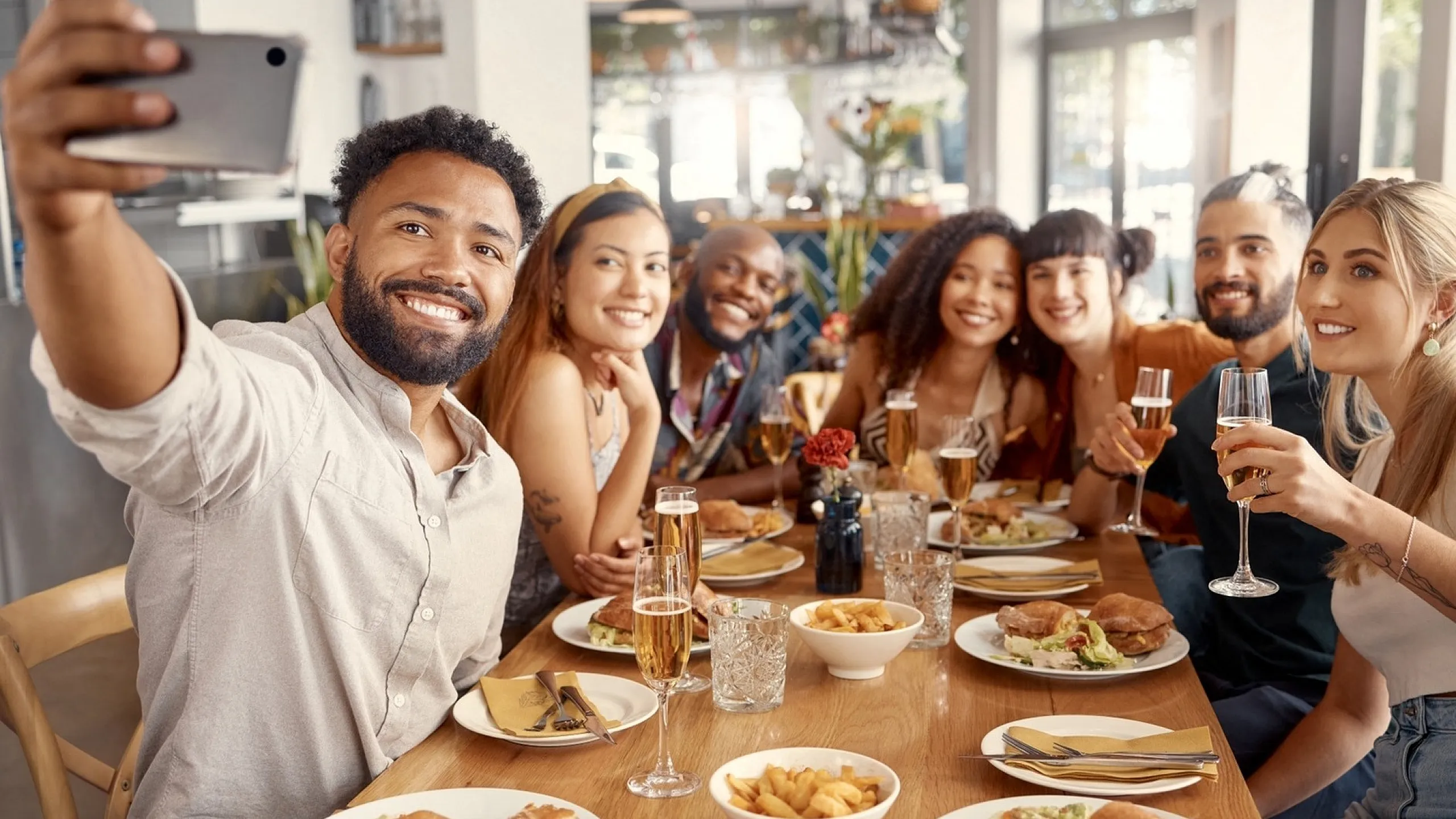 A group of friends at a restaurant table taking a selfie, with plates of food and drinks in front of them.