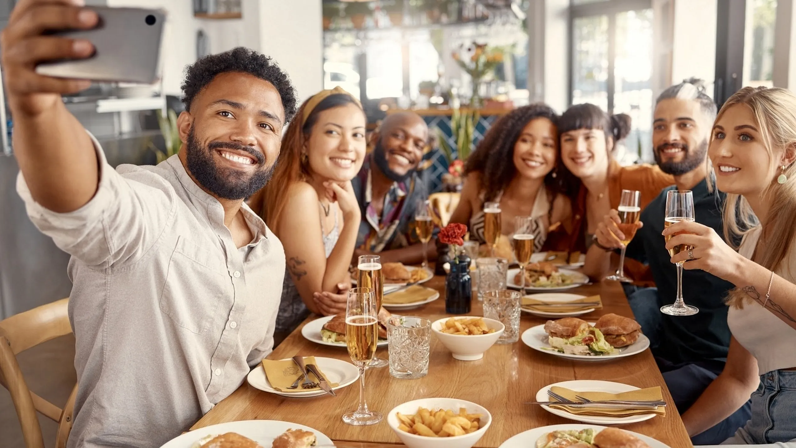 A group of friends at a restaurant table taking a selfie, with plates of food and drinks in front of them.