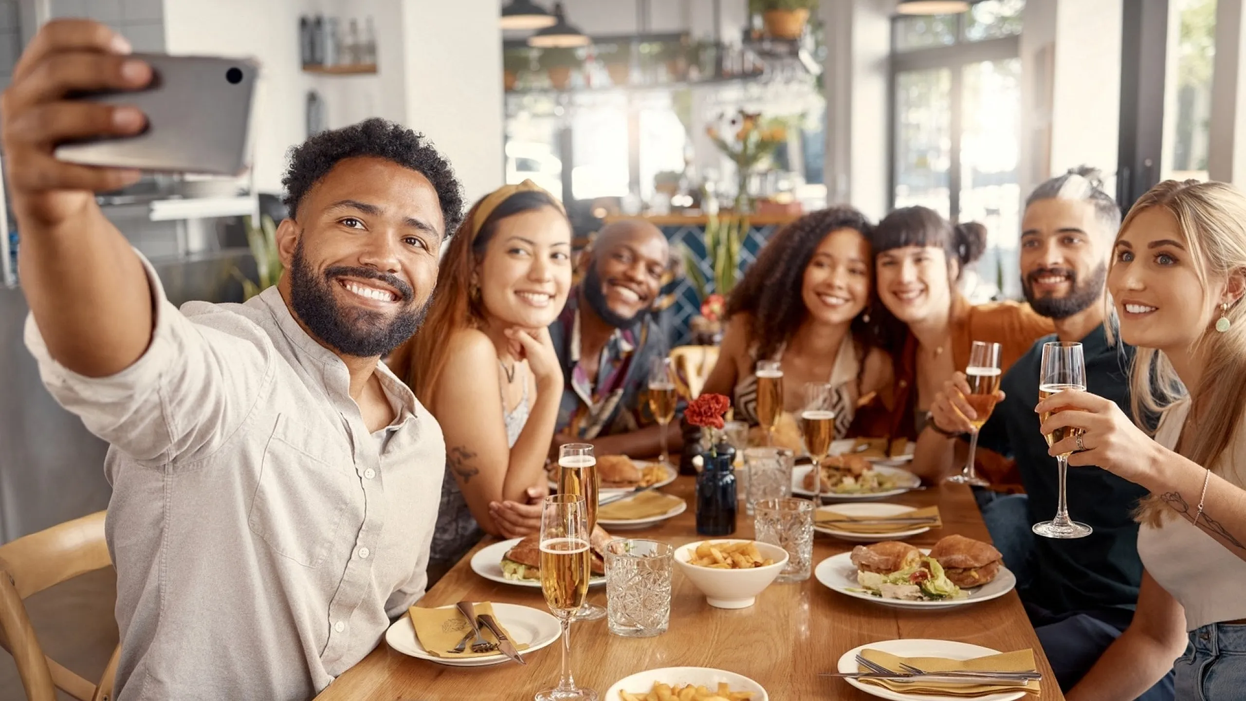 A group of friends at a restaurant table taking a selfie, with plates of food and drinks in front of them.
