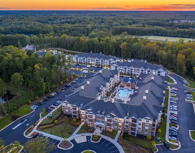 Elevated view of a modern apartment complex with a stunning sunset backdrop, featuring beautifully landscaped grounds, ample parking, and a central pool area surrounded by lush forest.