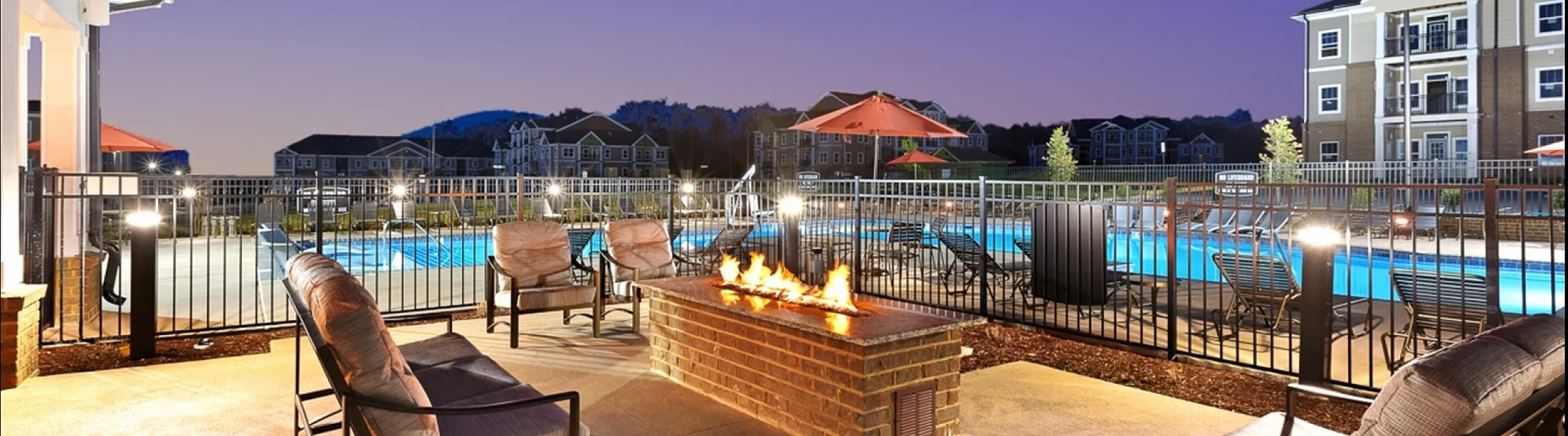 Chic outdoor lounge with cozy firepit, overlooking a sparkling pool and stylish apartments at dusk.