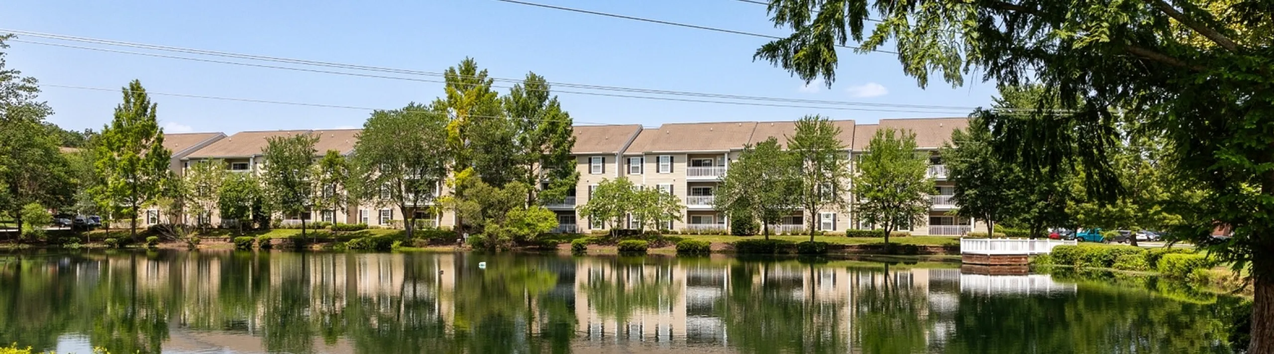 Serene lakeside view of an apartment complex surrounded by lush greenery and clear blue skies.