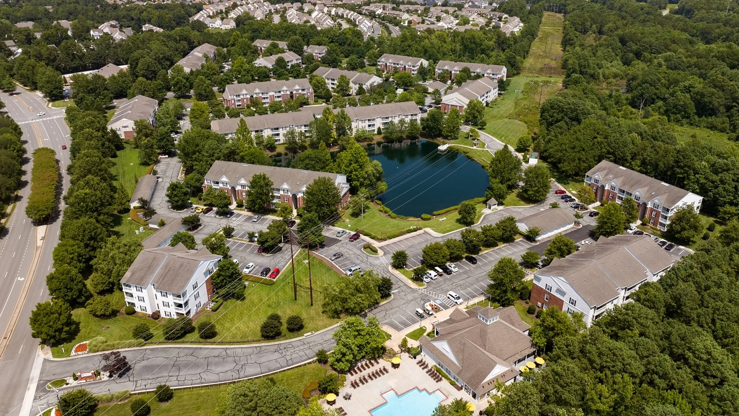 Aerial view of a lush apartment community surrounded by green landscapes, featuring a tranquil pond and a sparkling swimming pool.