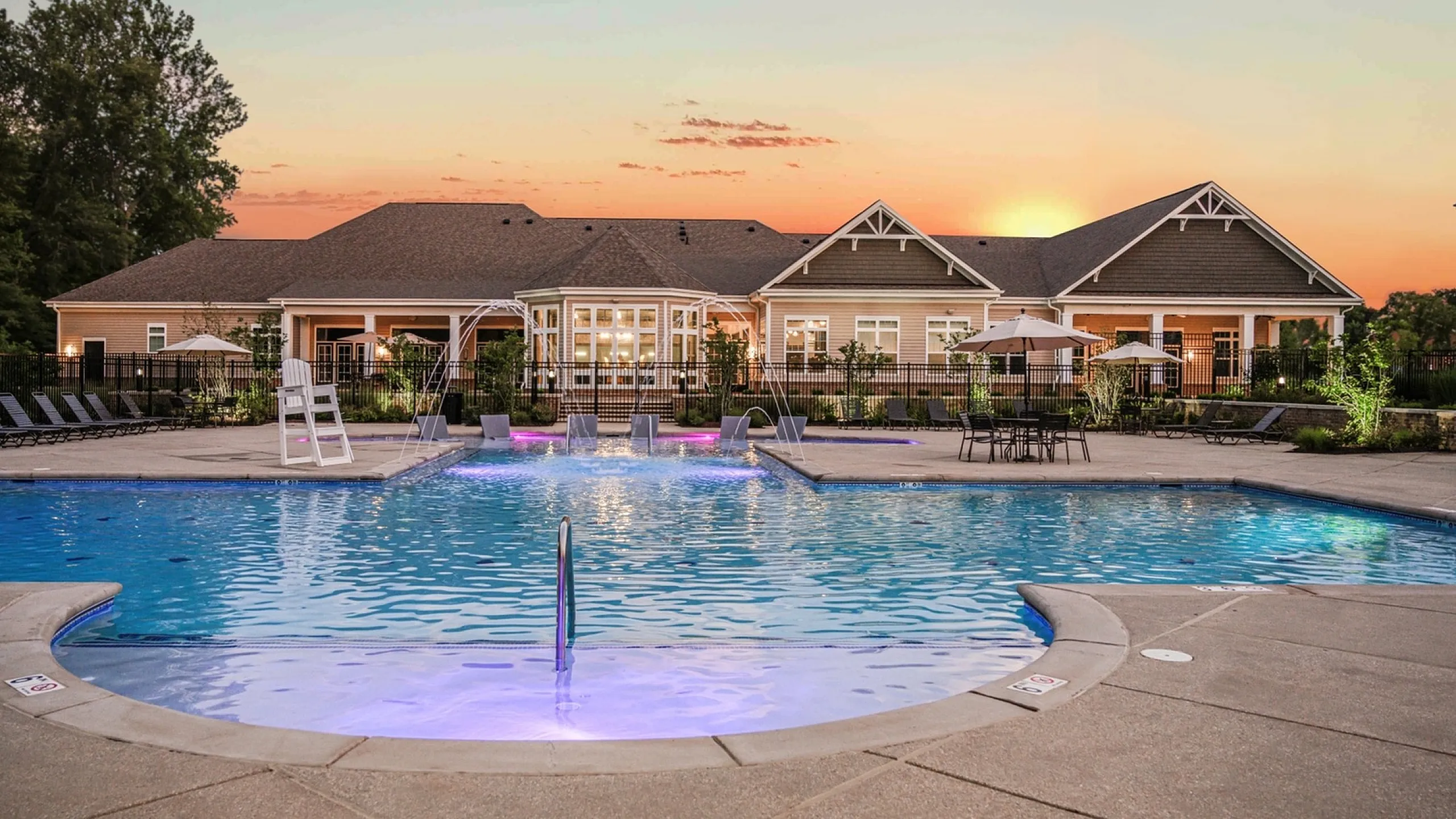 Luxurious apartment complex featuring a stunning illuminated pool area with stylish lounge chairs, surrounded by elegant architecture at sunset.