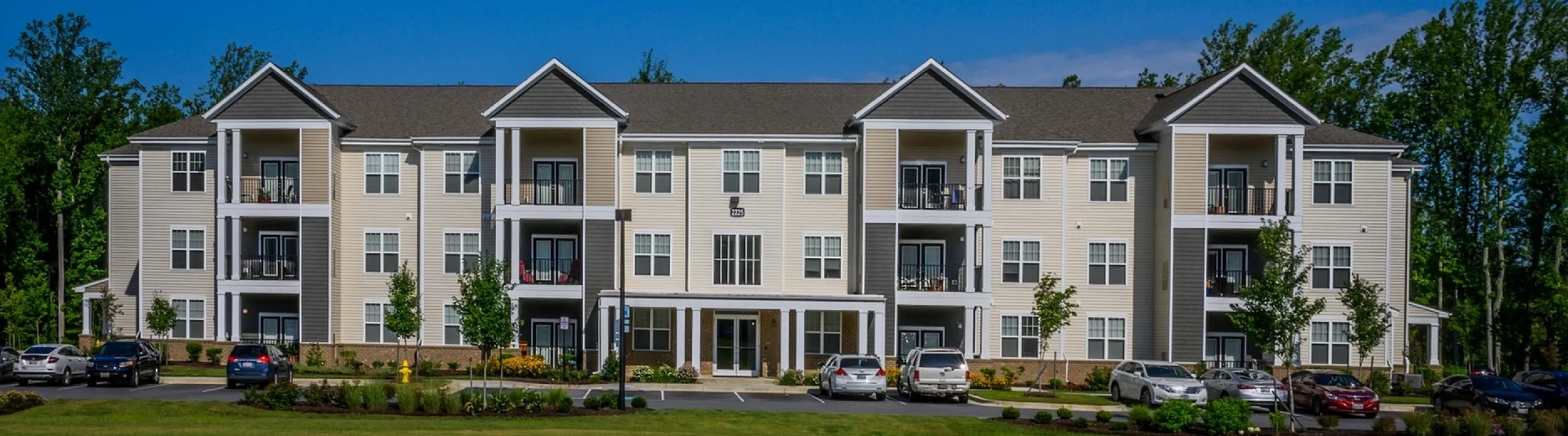 Modern three-story apartment building with balconies, surrounded by lush greenery and a spacious lawn, under a clear blue sky.