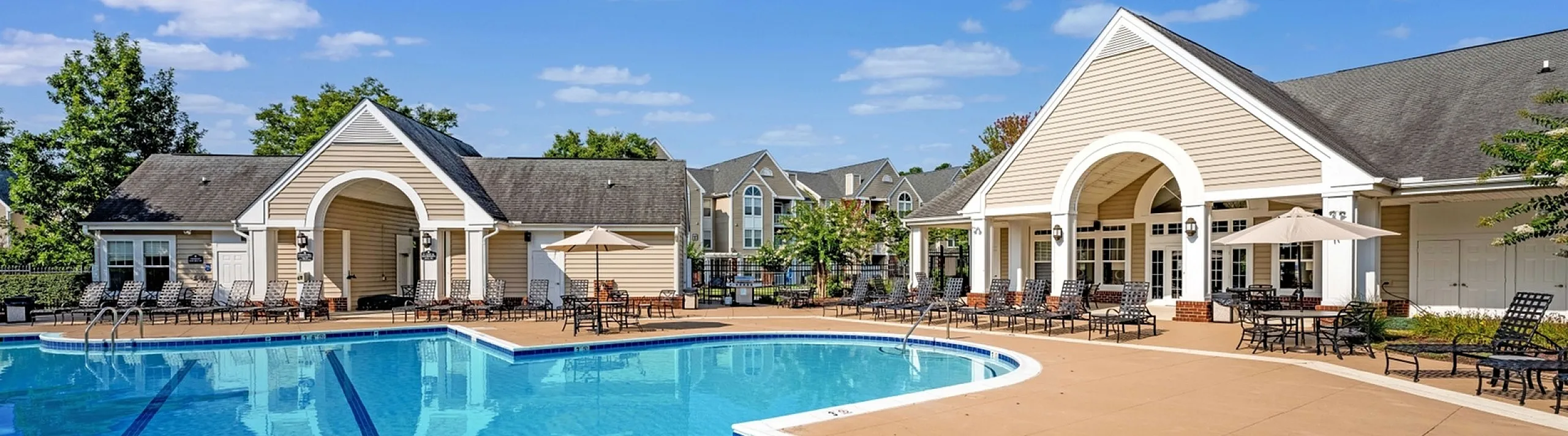 Inviting apartment community pool with elegant sun loungers and shaded seating areas, nestled under a clear blue sky.