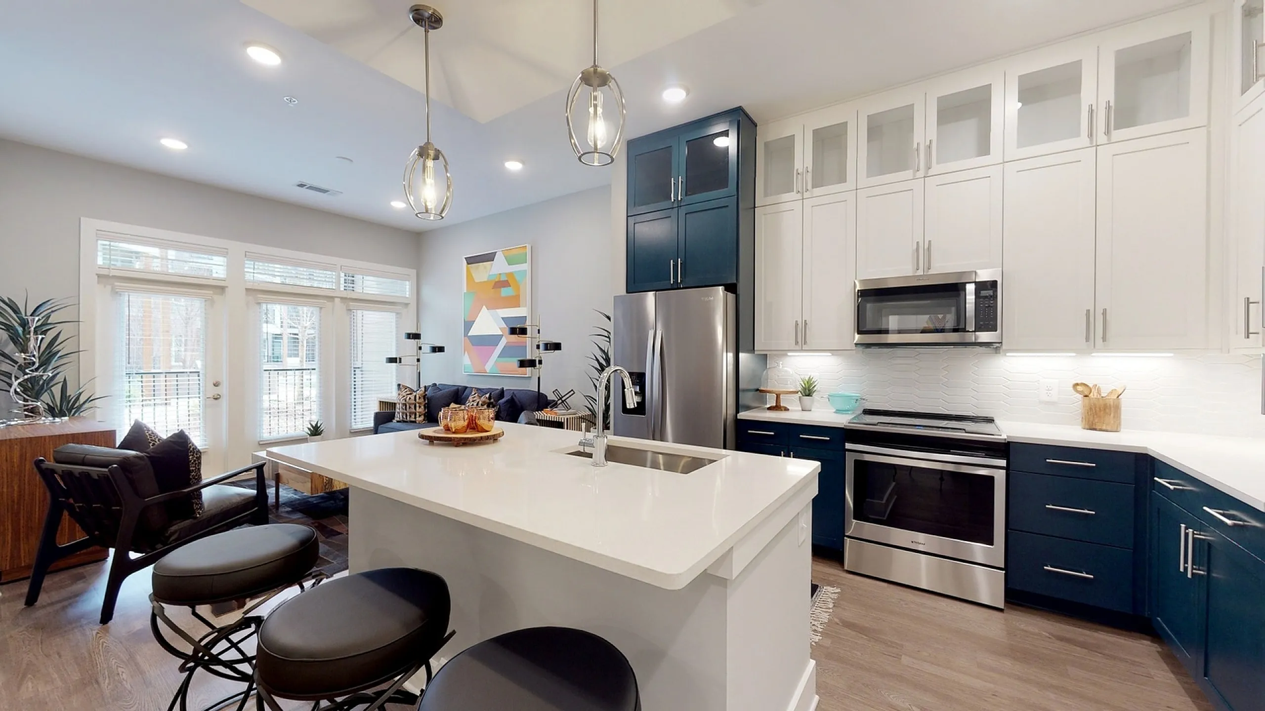 Contemporary open-plan kitchen with sleek white countertops, stainless steel appliances, and elegant lighting, flowing into a stylish living area with large windows and modern decor.