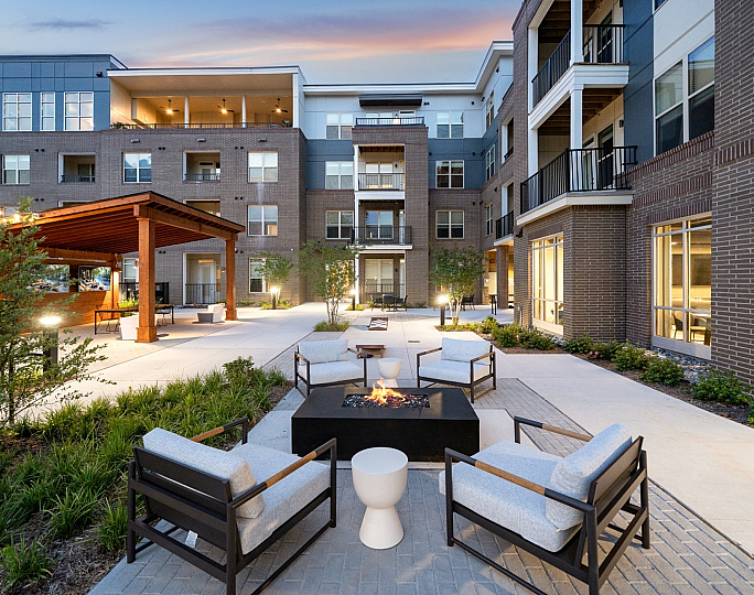 Modern apartment complex courtyard at sunset featuring cozy outdoor seating around a fire pit with stylish wooden pergola and lush landscaping.