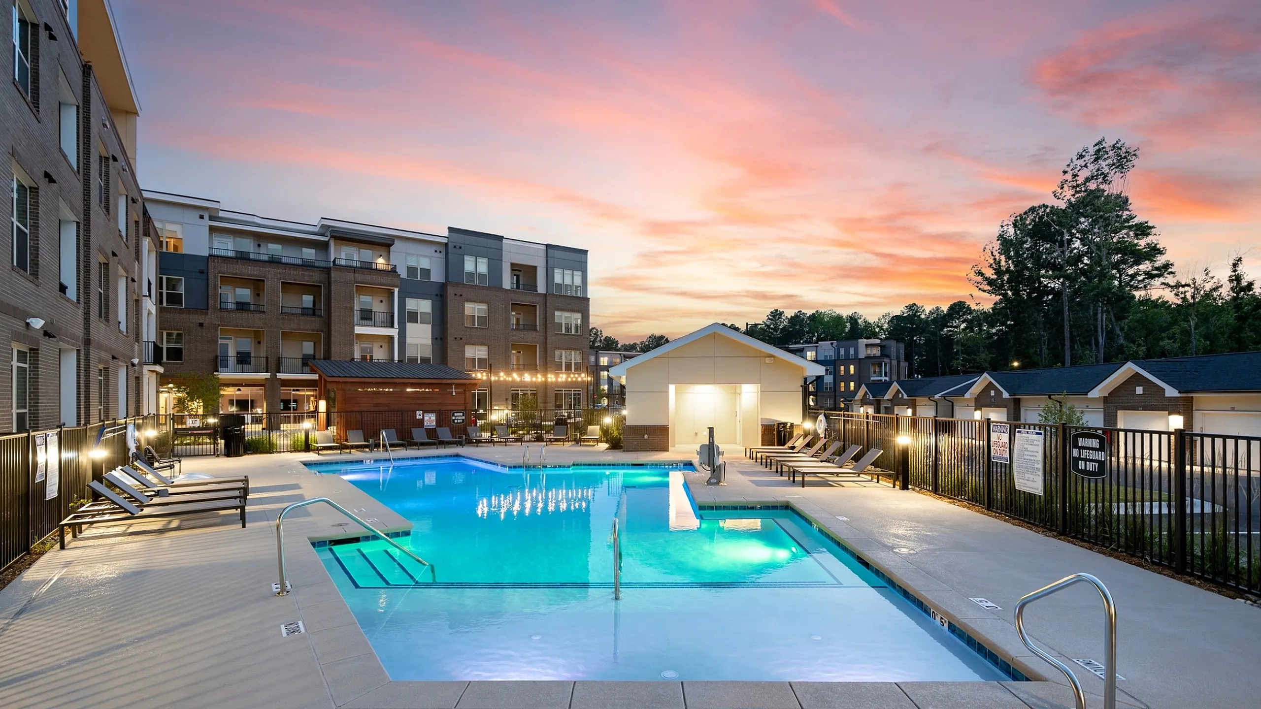 Luxurious apartment complex with a stunning pool area, surrounded by modern architecture and highlighted by a vibrant sunset sky.