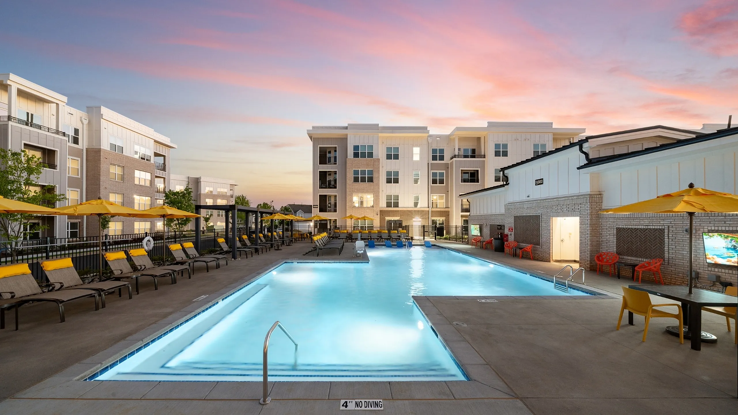 Luxurious apartment complex with a stunning outdoor pool and sunset views, featuring modern architecture and inviting lounge chairs with vibrant yellow umbrellas.