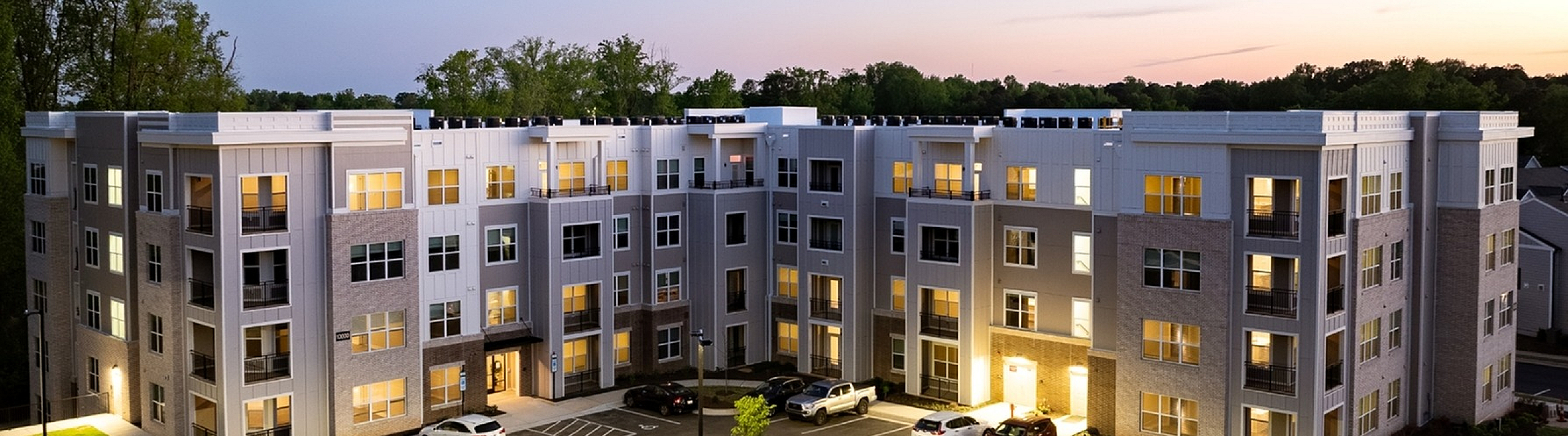 Modern apartment complex at dusk with elegant lighting, ample parking, and lush greenery.