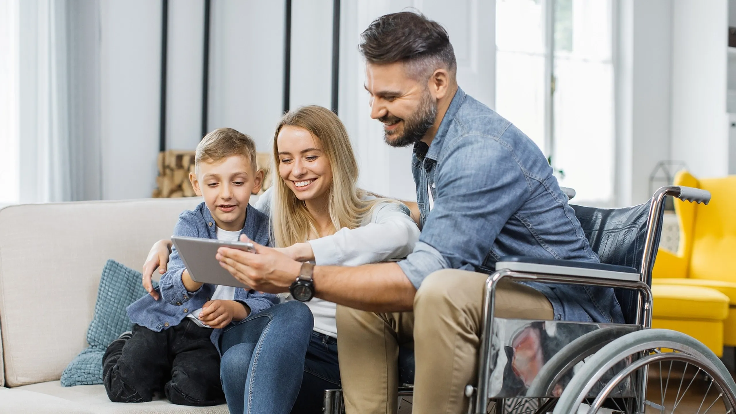 Happy family in a modern living room, a father in a wheelchair, mother, and son smiling while looking at a tablet, showcasing an inclusive and welcoming apartment space.