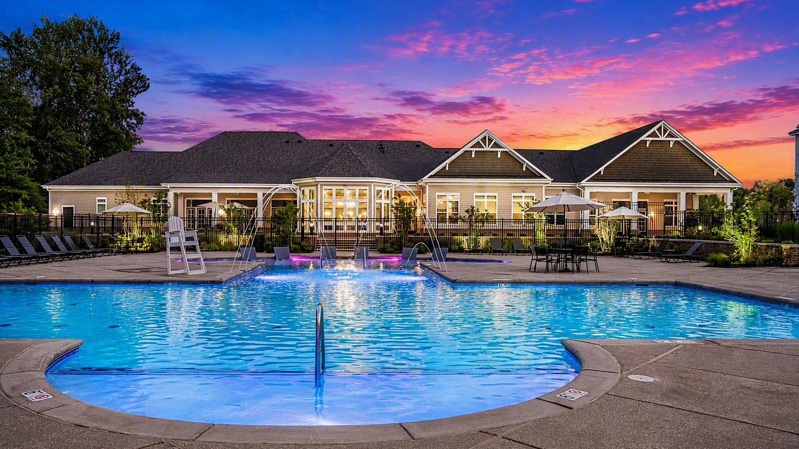 Luxurious apartment community with a stunning illuminated swimming pool, surrounded by elegant poolside loungers and vibrant sunset sky.