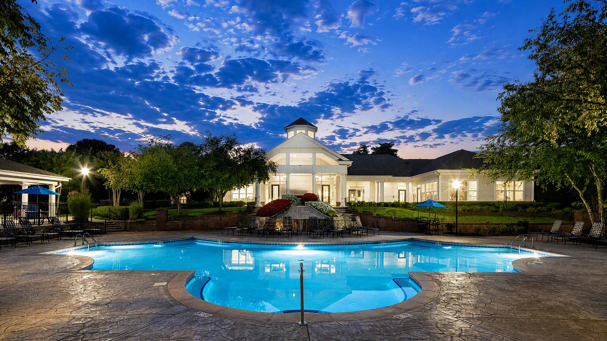 Luxurious apartment complex with a sparkling blue pool, surrounded by lush gardens and elegant architecture under a picturesque twilight sky.