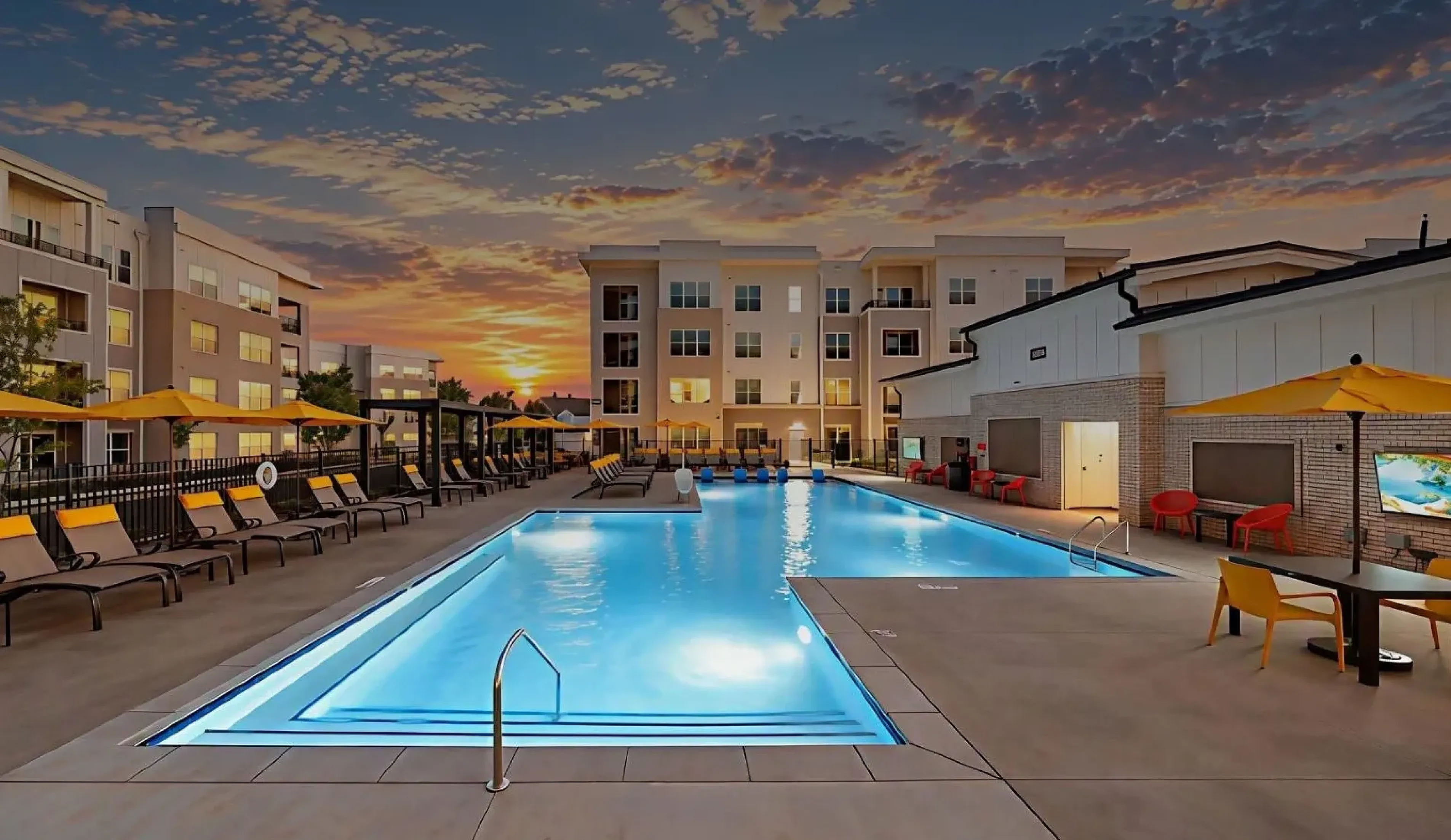 Resort-style outdoor swimming pool at Abberly Commons in Raleigh, NC, featuring illuminated blue water, spacious sun deck seating, yellow umbrellas and contemporary apartment buildings surrounding the courtyard at sunset.