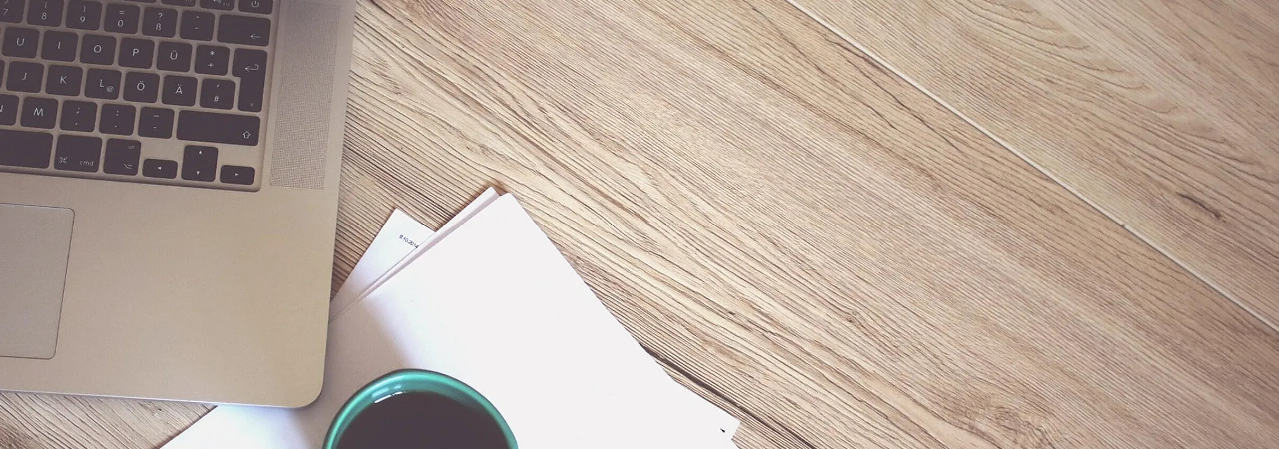 Modern workspace with sleek laptop, fresh coffee in a teal mug, and minimalistic papers on elegant wooden desk.
