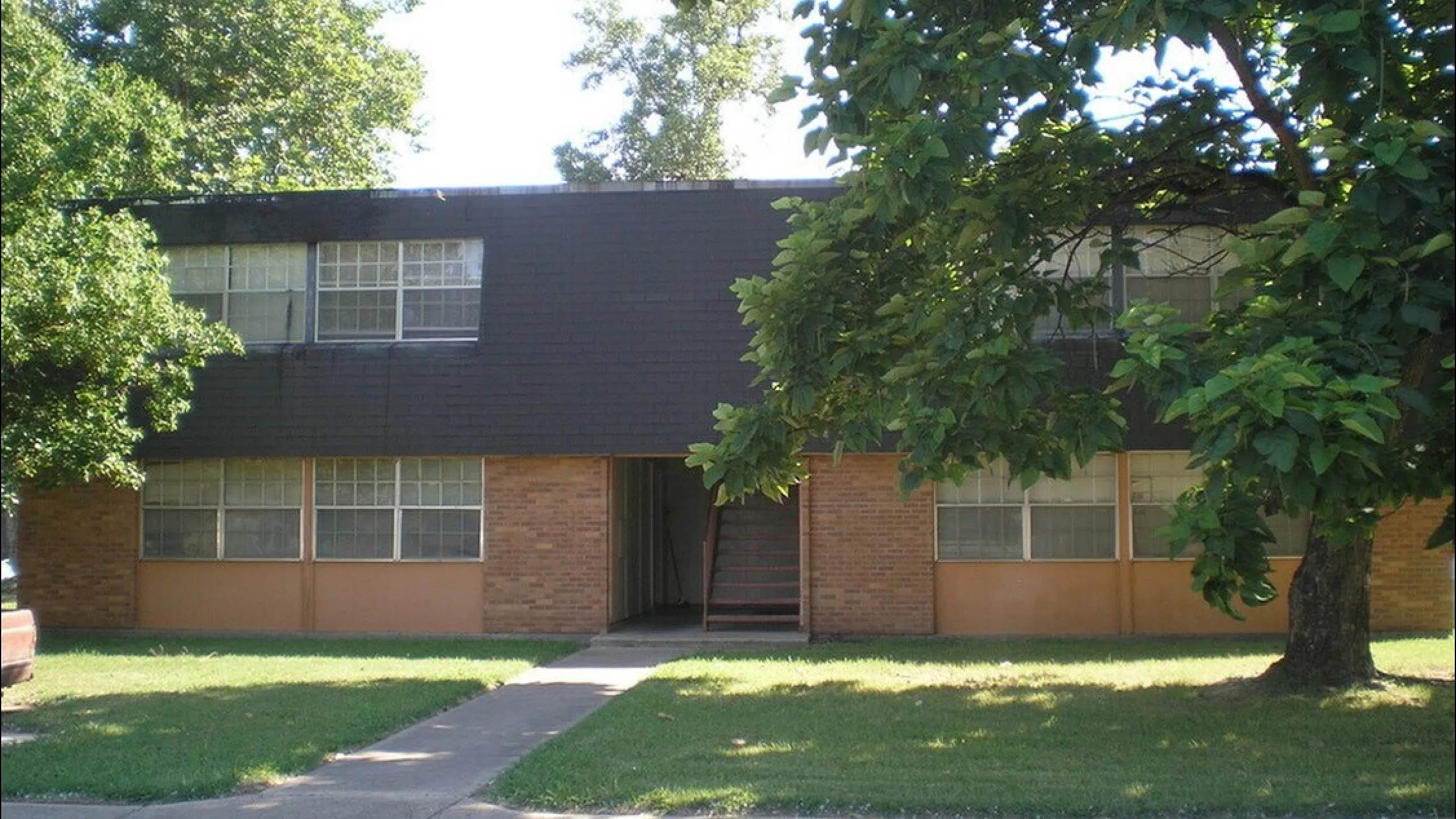 Charming two-story brick apartment building surrounded by lush greenery and mature trees, offering a serene residential setting.