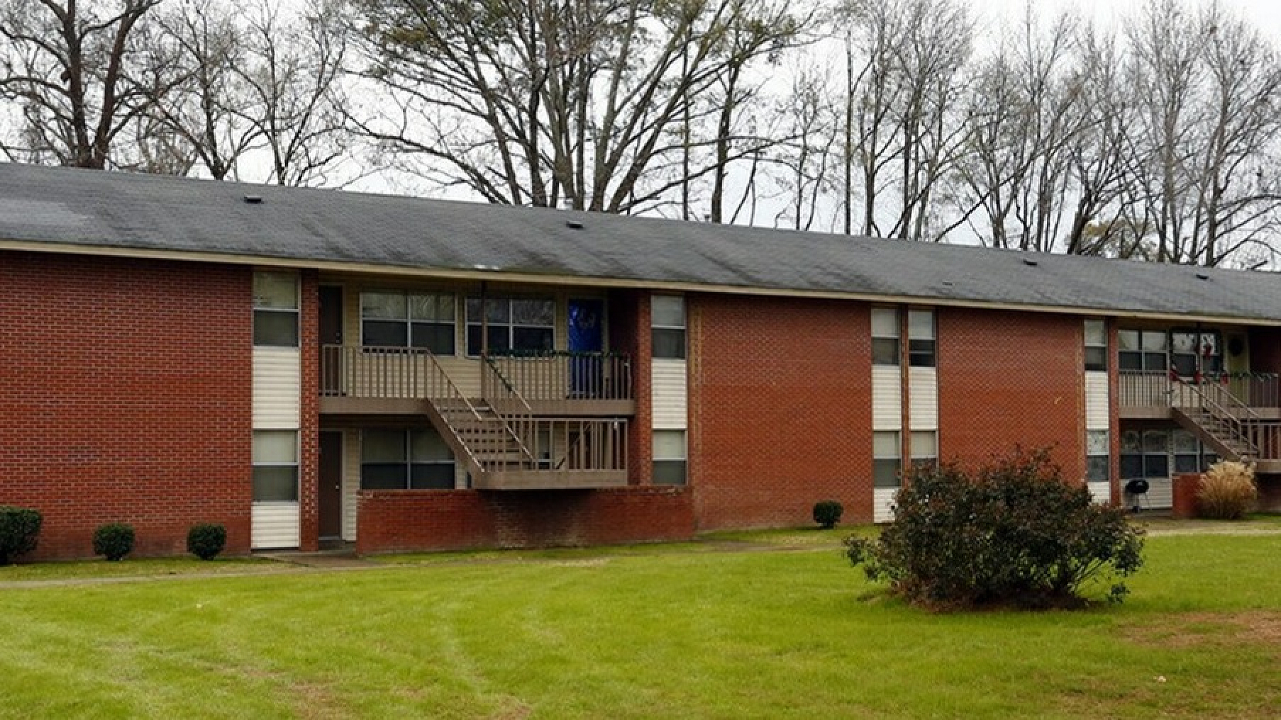 Charming brick apartment building with lush green lawns, surrounded by serene wooded area and inviting balconies.