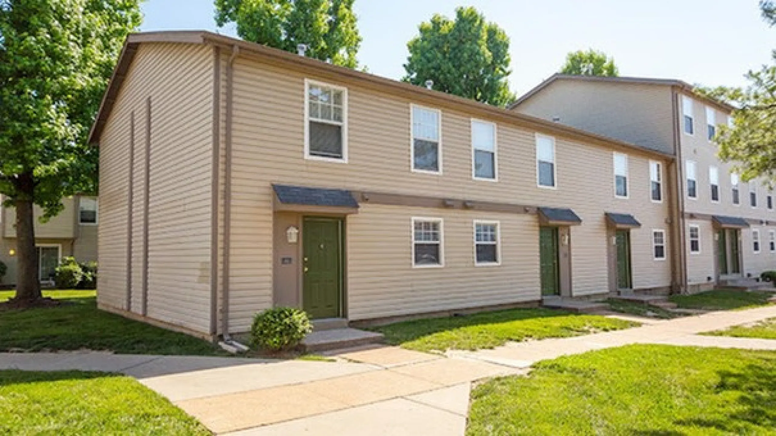 Charming apartment building with beige siding surrounded by lush greenery and bright pathways in a peaceful neighborhood.