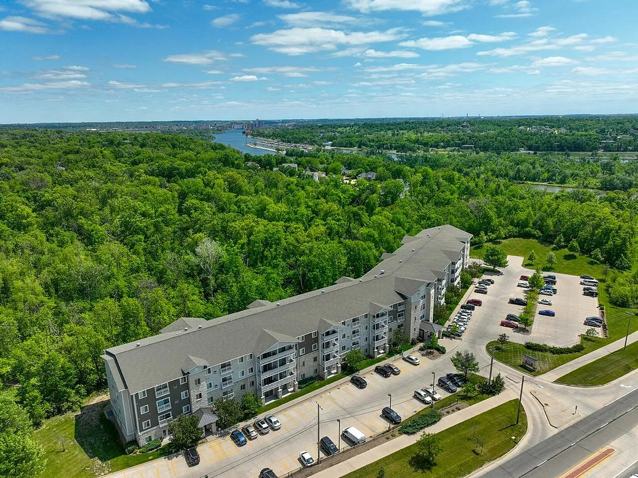 Scenic aerial view of a modern riverside apartment complex surrounded by lush greenery, offering tranquil living with ample parking and easy access to nature.