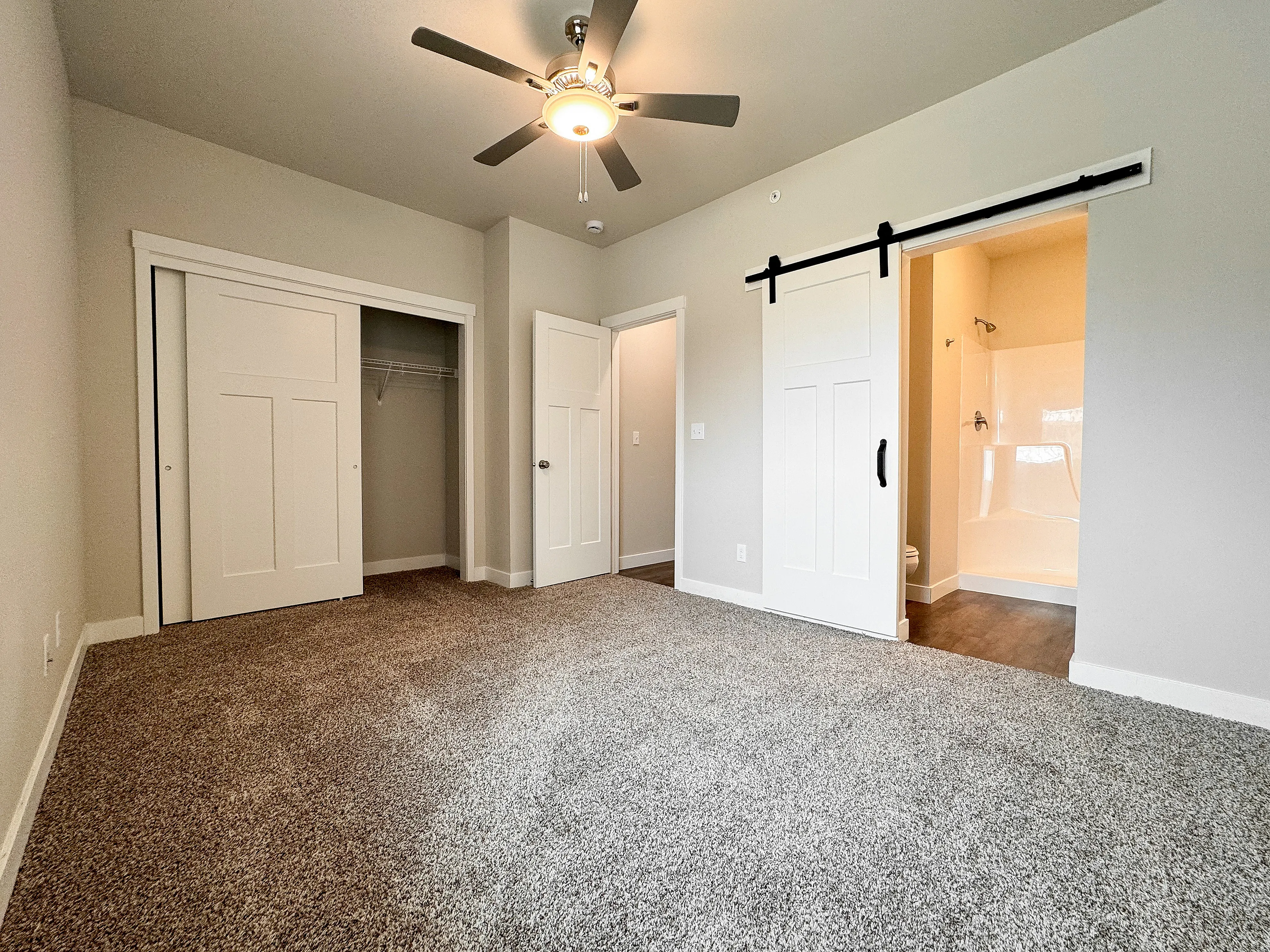 Spacious bedroom featuring plush carpeting, a sleek ceiling fan, and a modern barn door leading to an elegant en-suite bathroom.