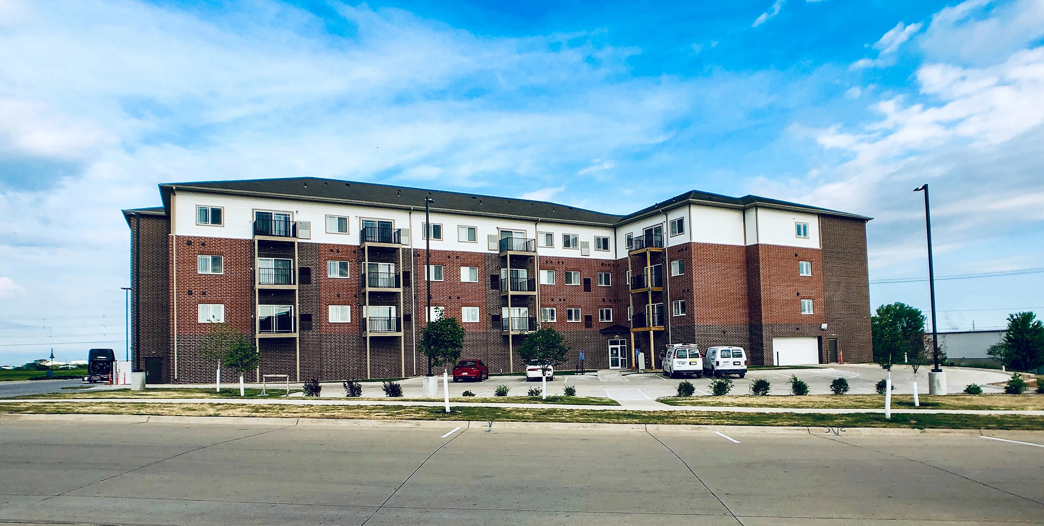 Modern brick apartment building with balconies, surrounded by a well-maintained parking area and landscaped greenery under a vibrant blue sky.