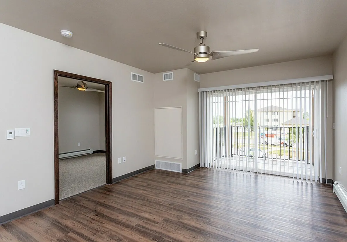 Bright and airy apartment living room with modern ceiling fan, large windows, and wooden flooring, leading into a cozy bedroom.