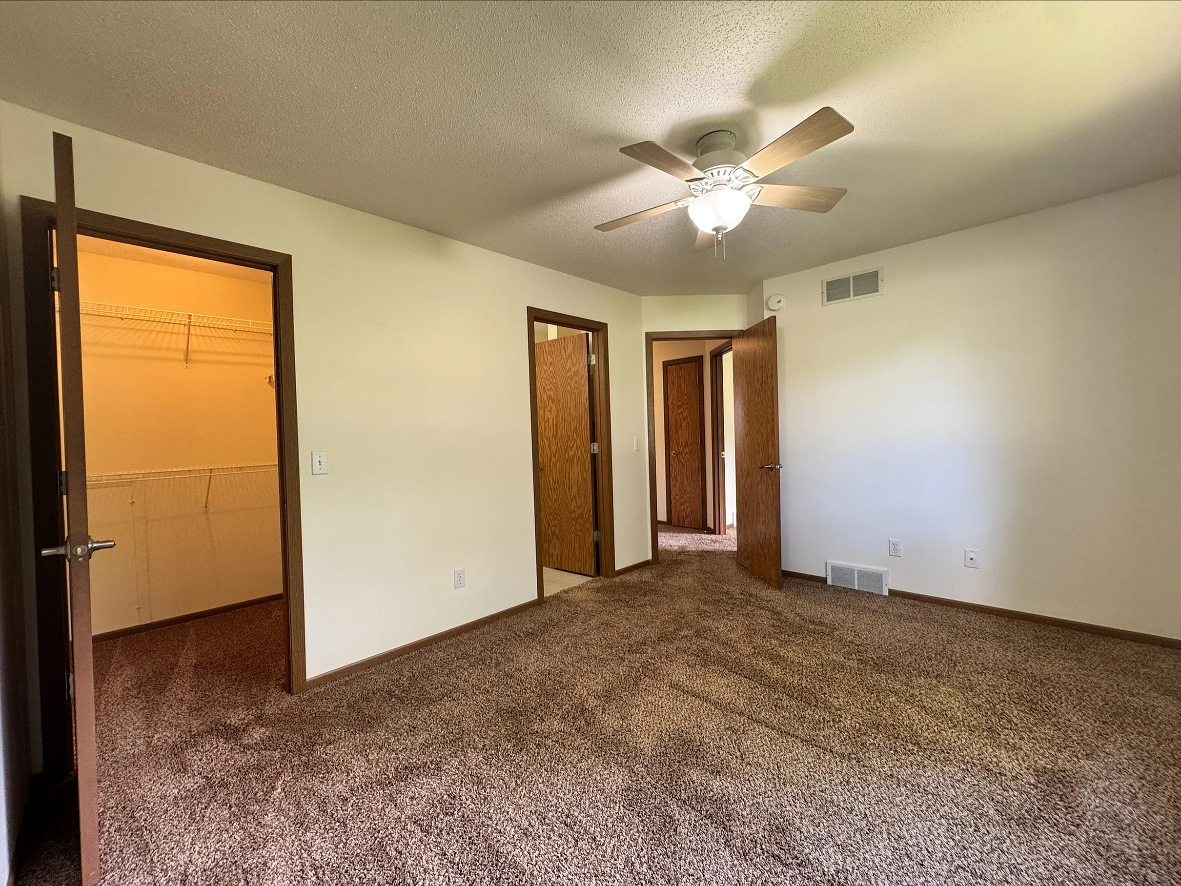 Bright bedroom with plush carpet, ceiling fan, and two doors leading to spacious walk-in closets.
