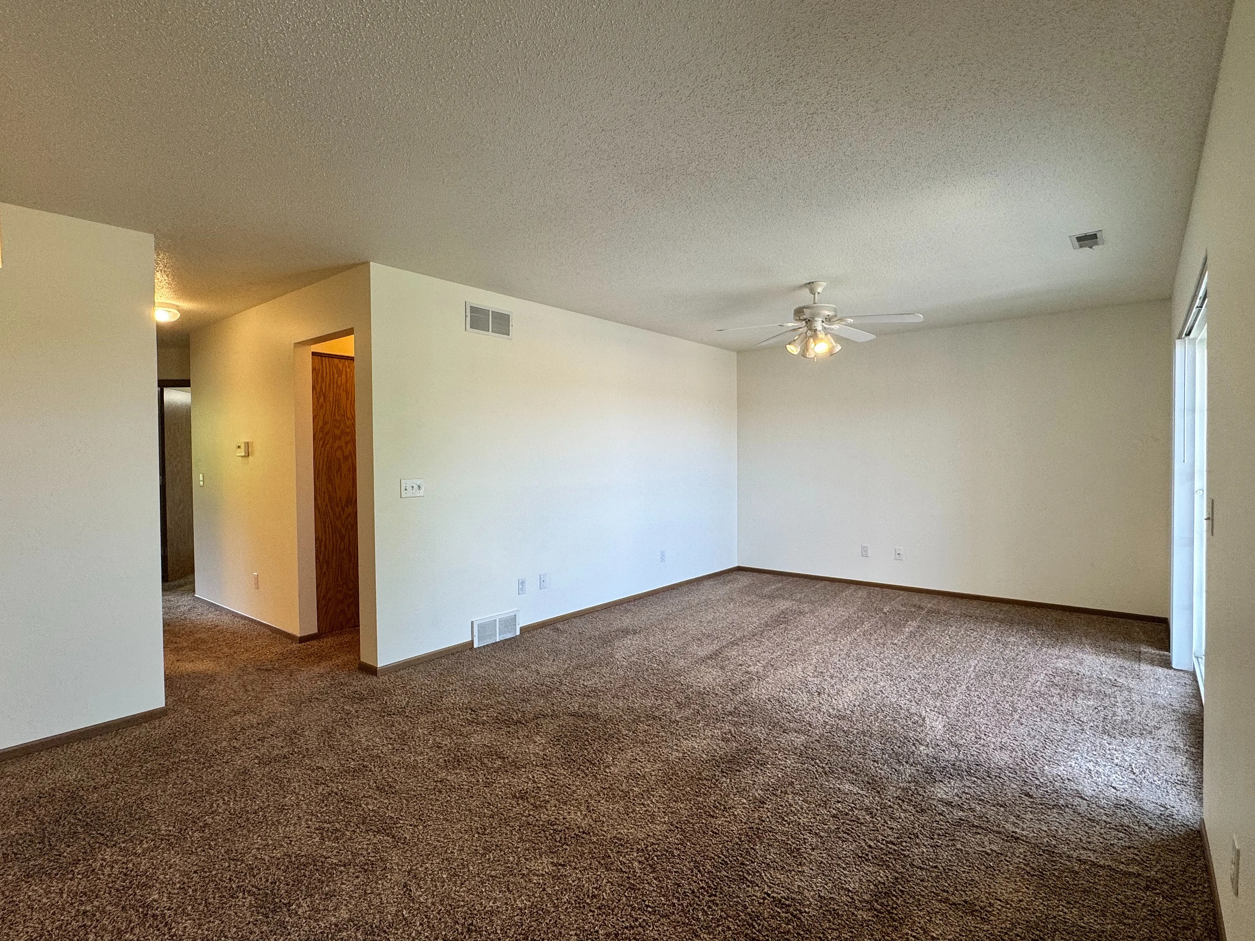 Spacious living room with plush brown carpeting, white walls, and ceiling fan, illuminated by natural light.