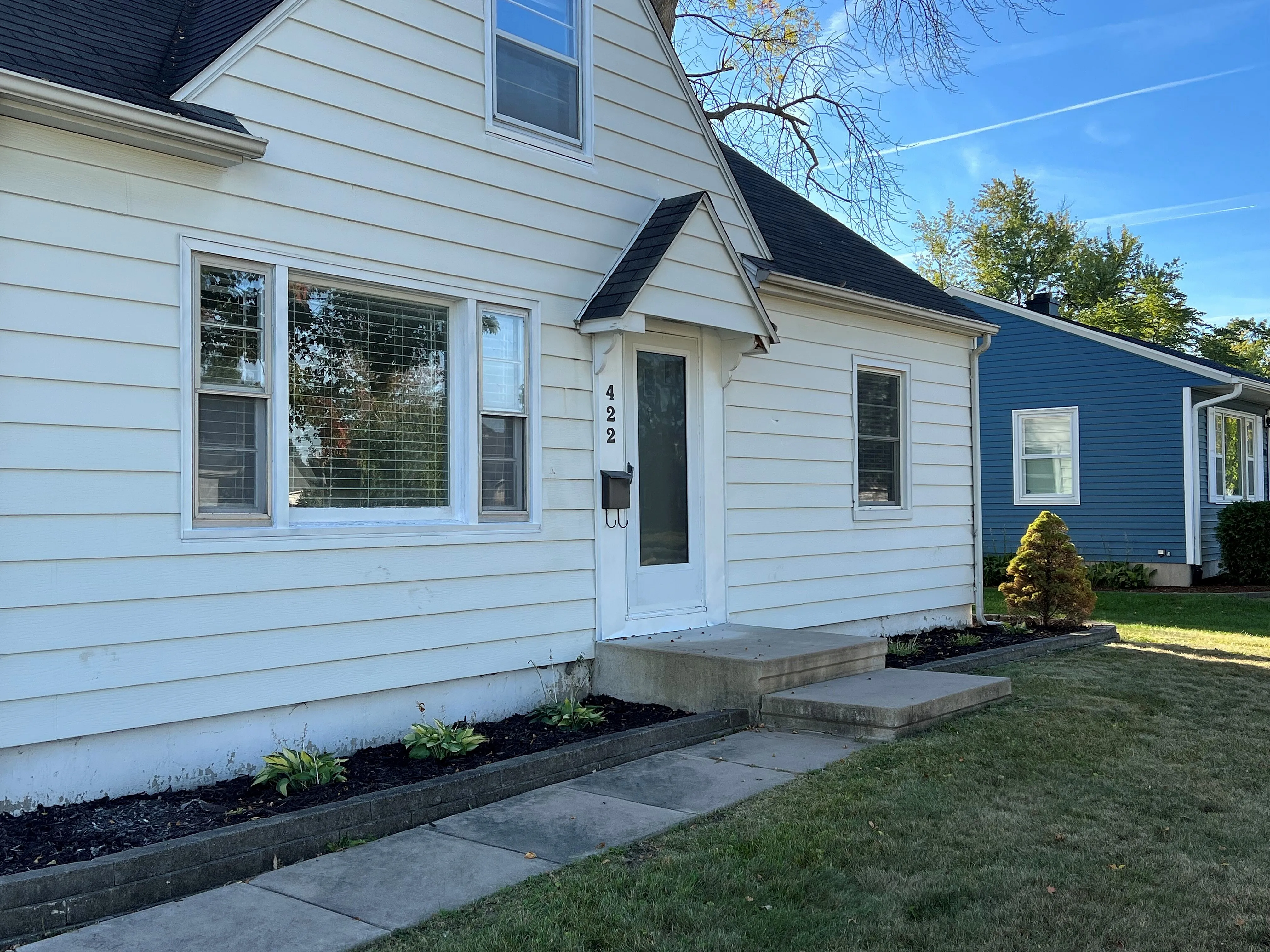 Charming single-story house with fresh white siding, large windows, and welcoming front steps, nestled in a serene neighborhood with lush green lawns.