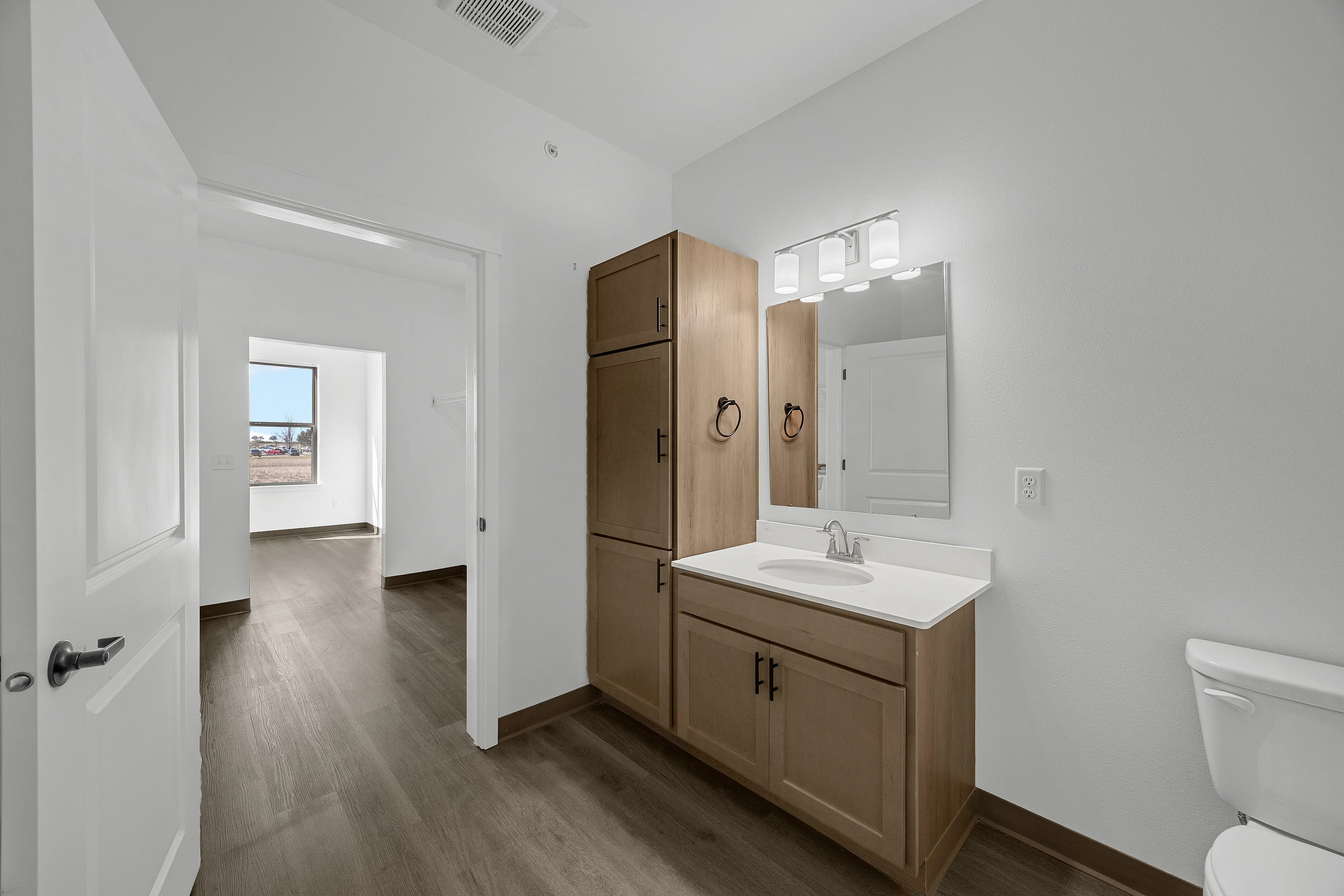Sleek and modern bathroom with wooden cabinets, elegant lighting, and a view into a sunlit room with stylish wood flooring.