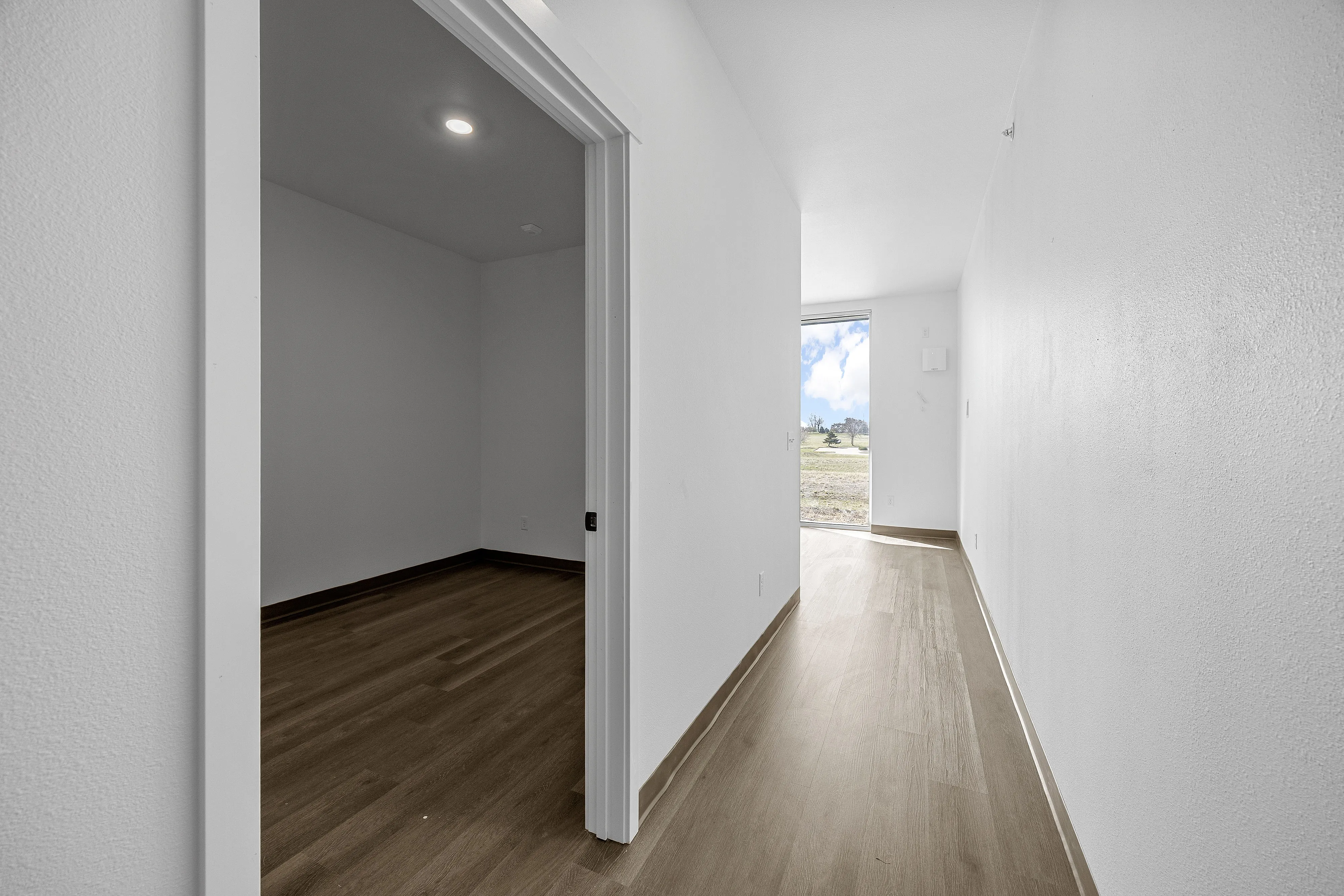 Bright apartment hallway with sleek wooden flooring leading to a sunlit room with expansive outdoor views.