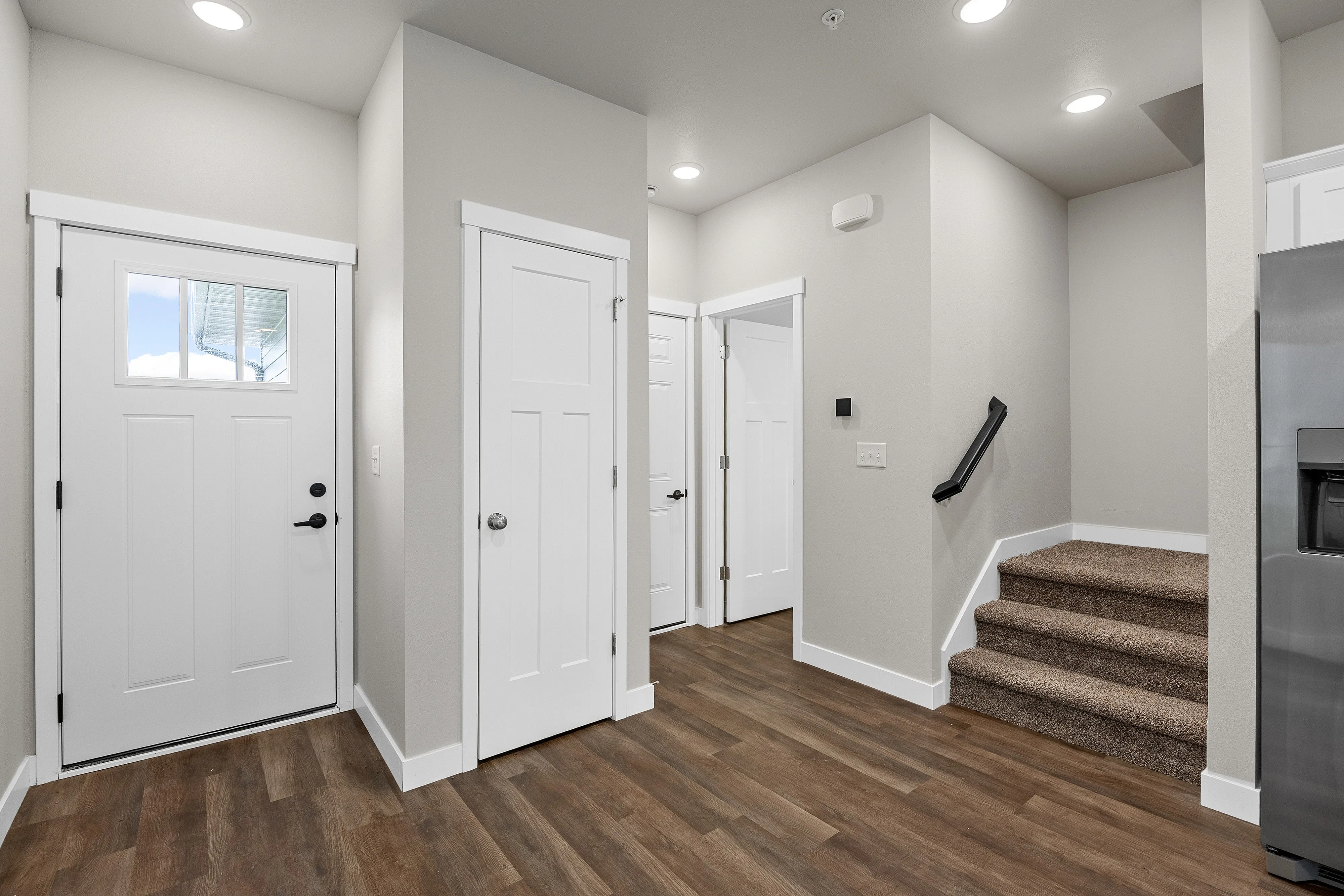 Contemporary foyer with warm wood flooring, sleek white doors, and modern recessed lighting, leading to a cozy carpeted staircase.