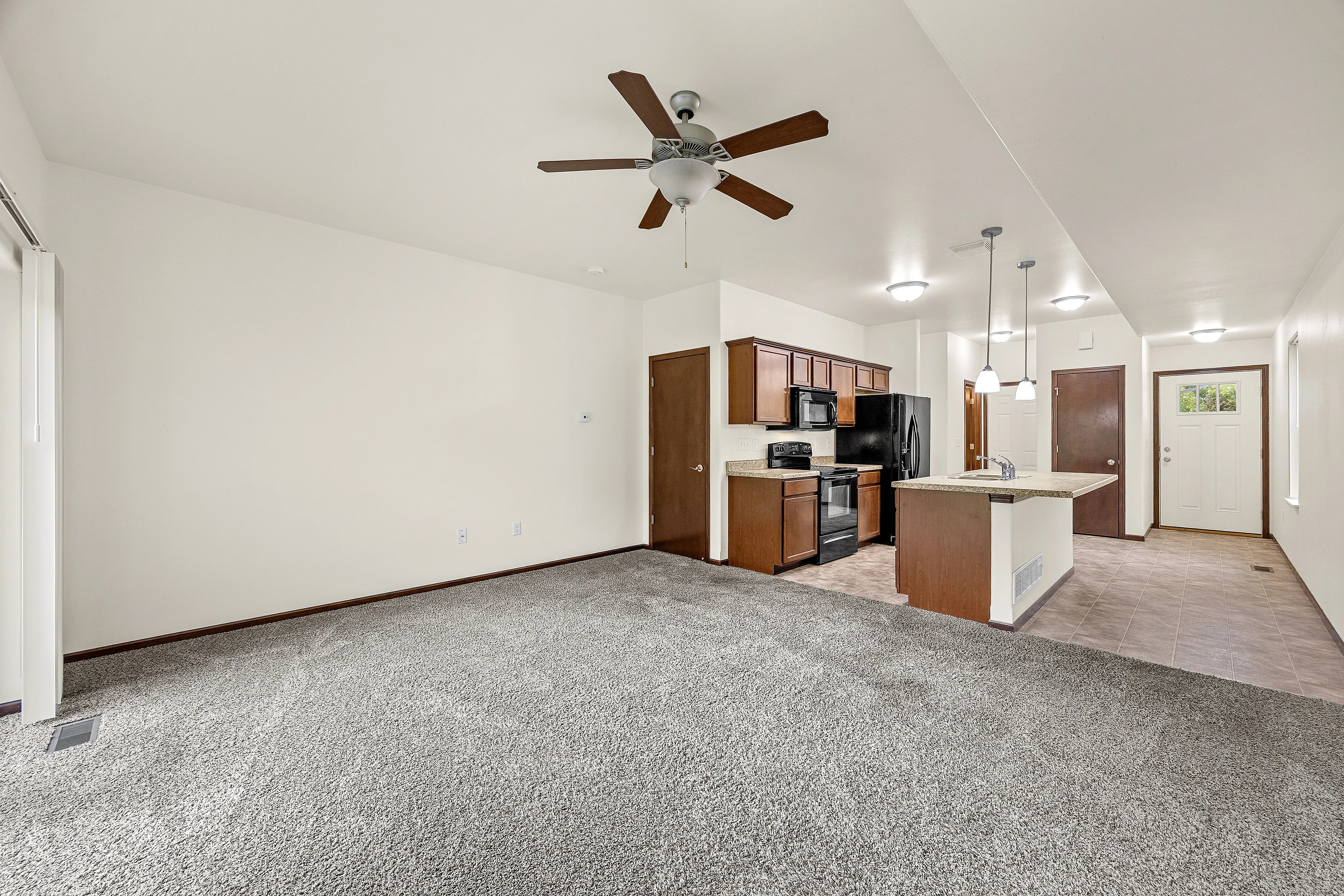 Modern open-concept living space with plush carpeting, ceiling fan, and contemporary kitchen featuring sleek appliances and elegant cabinetry.