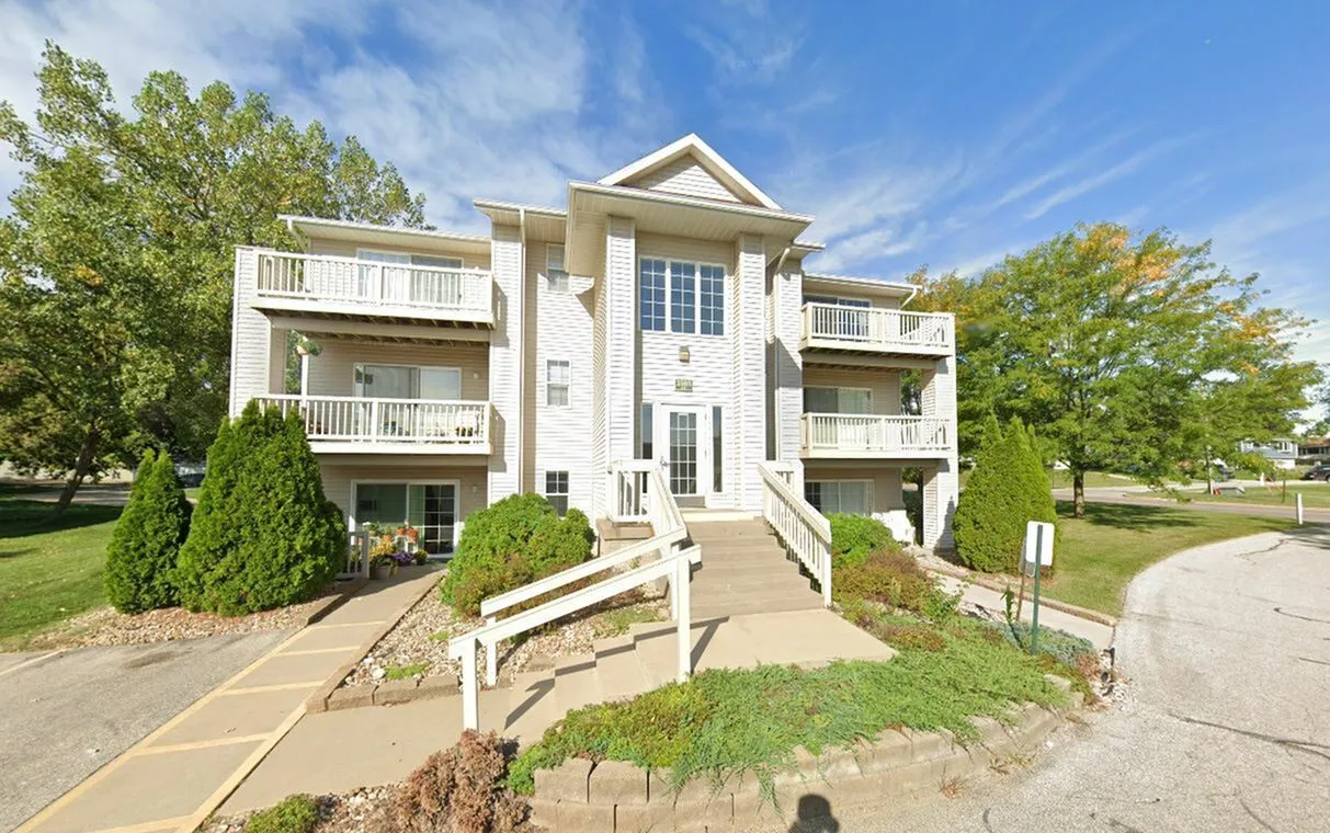 Charming three-story white apartment building with spacious balconies, surrounded by lush green trees and a clear blue sky.