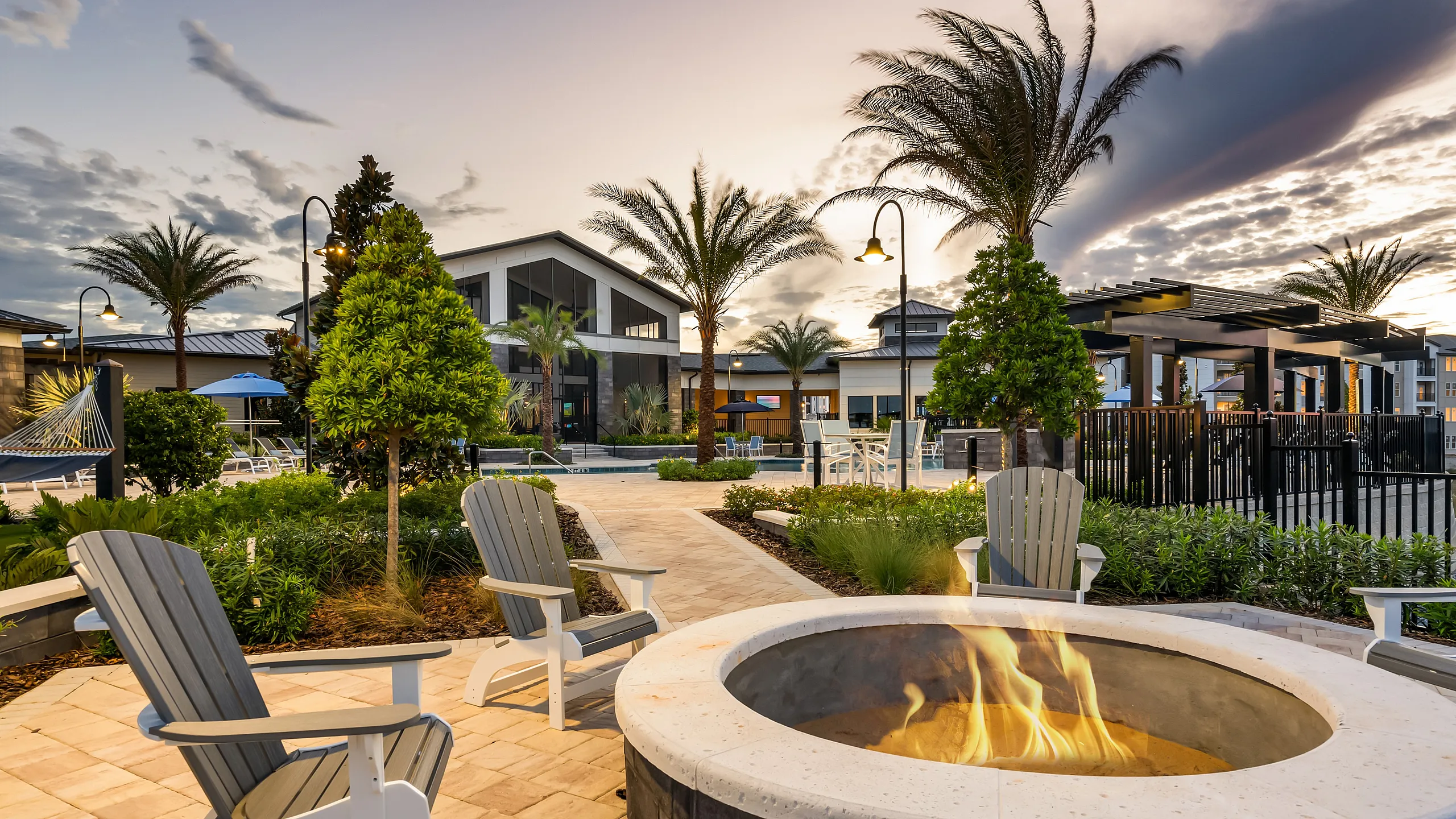 Luxurious outdoor oasis with fire pit, palm trees, and elegant seating overlooking a modern community setting at sunset.