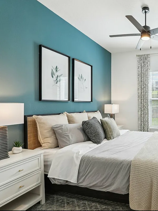 Elegant bedroom with a modern ceiling fan, cozy bedding, and stylish artwork on a teal accent wall, complemented by natural light through a large window.