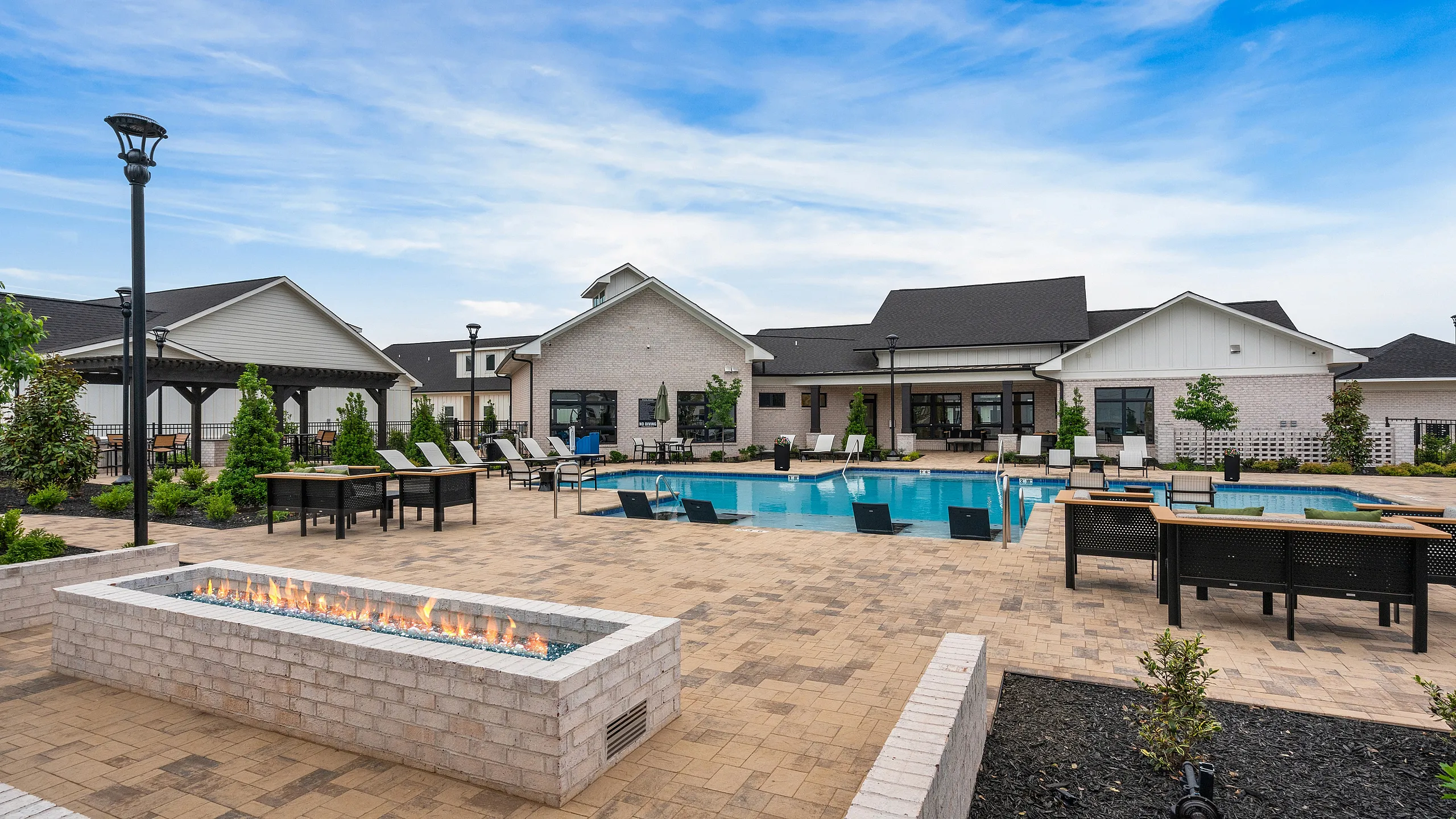 Luxurious apartment complex featuring a sparkling pool, modern lounge chairs, and an elegant outdoor fire pit area against a serene blue sky backdrop.