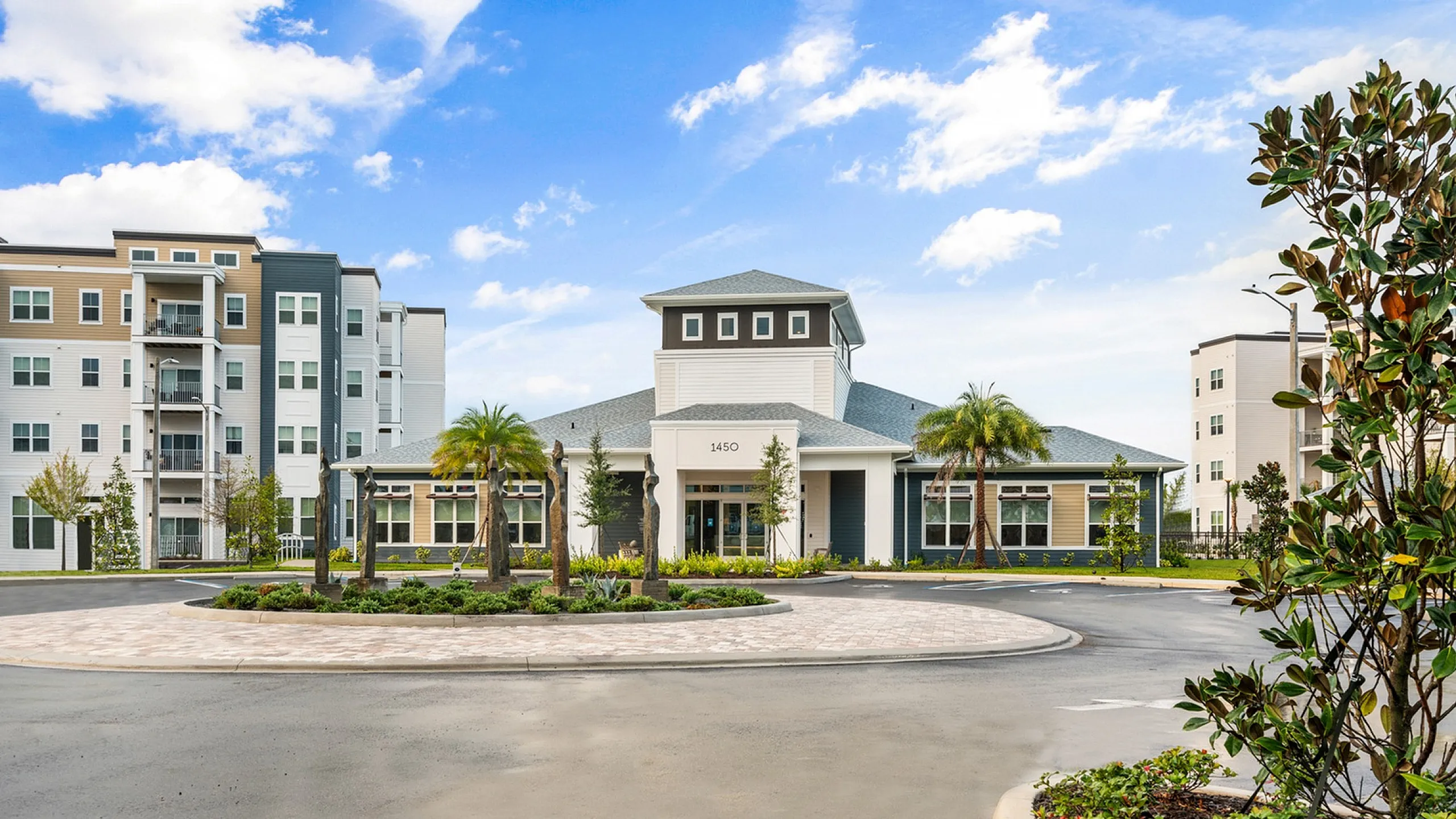 Modern apartment complex with lush landscaping, palm trees, and contemporary architecture under a vibrant blue sky.